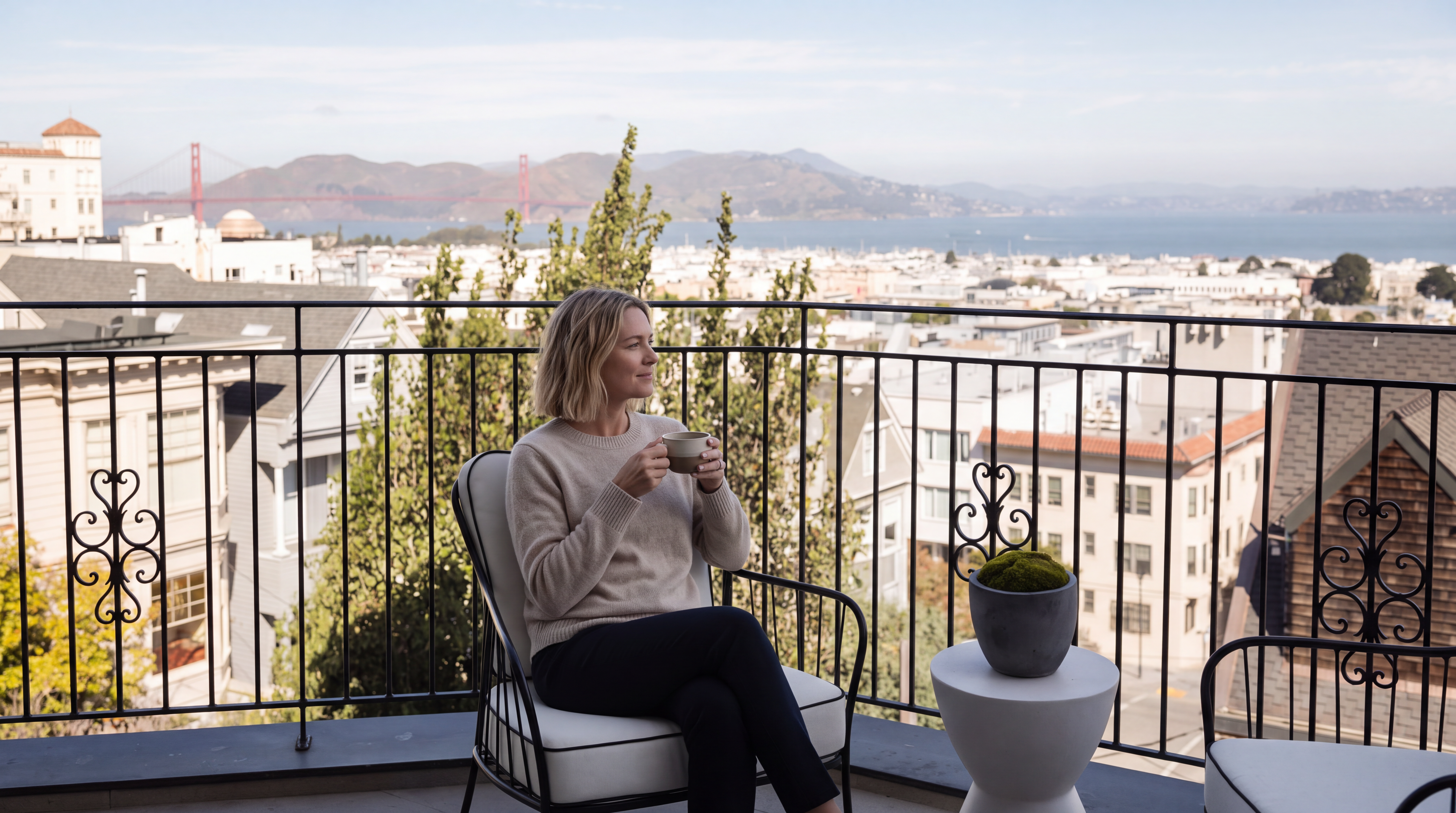 This elevated balcony offers a stunning, panoramic view of the San Francisco skyline, including the iconic Golden Gate Bridge in the distance. The space is furnished with two modern, white-cushioned chairs and a minimalist white pedestal table topped with a small potted plant. The wrought-iron railing provides a secure perimeter while maintaining an open, airy feel, perfect for enjoying the urban scenery.