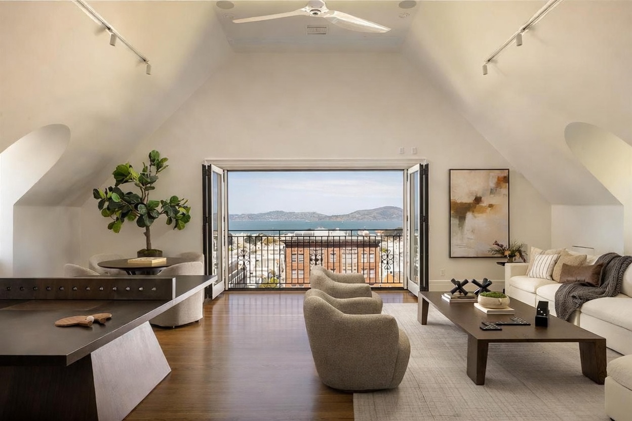 This sophisticated living room features a vaulted ceiling and an open-concept layout that seamlessly integrates a game area with a ping-pong table and a comfortable lounge space. Large folding glass doors open to a balcony, framing a stunning panoramic view of the bay and city skyline. The room is decorated with neutral-toned furniture, a textured area rug, and contemporary artwork, creating an airy and luxurious atmosphere.