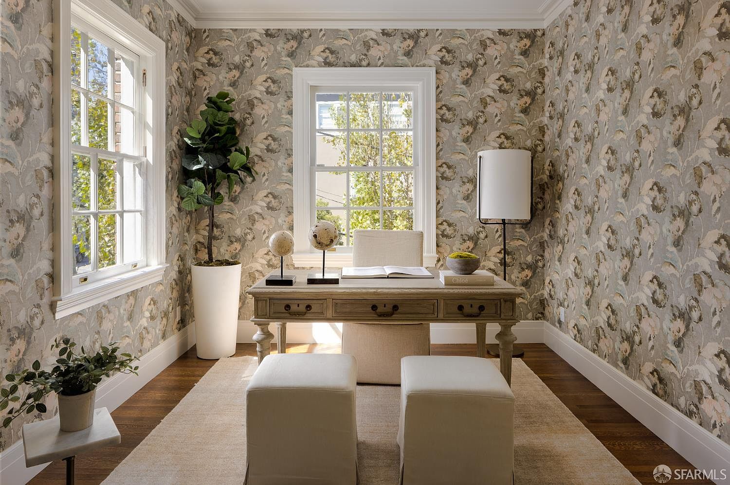 This elegant home office features sophisticated floral wallpaper and a central wooden desk paired with a matching upholstered chair. Two matching ottomans are positioned in front of the desk, while a tall potted plant and a floor lamp add warmth to the symmetrical layout. The room is bathed in natural light from two large windows, creating a serene and productive atmosphere.