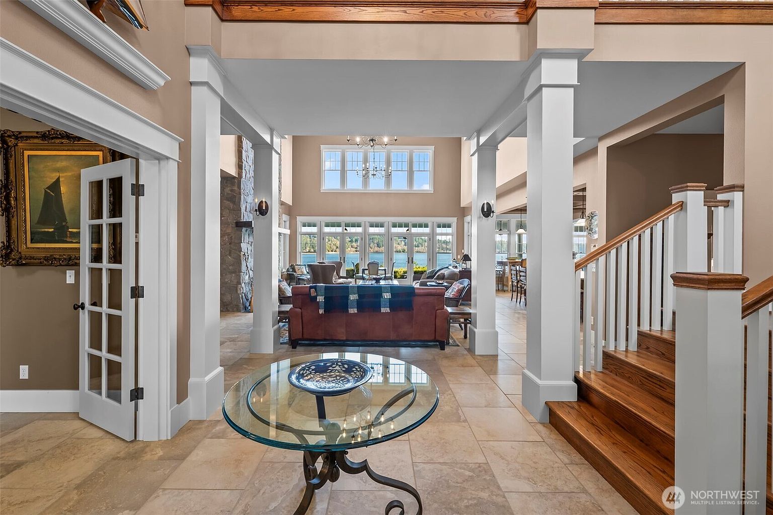 This grand entryway features a high-ceilinged foyer with a glass-topped pedestal table in the foreground and a staircase with wooden treads to the right. The open-concept layout provides a clear line of sight through the living area, which is anchored by a brown sofa and large windows overlooking a lake. The space is defined by neutral wall tones, tile flooring, and architectural columns that frame the transition between the entry and the main living space.