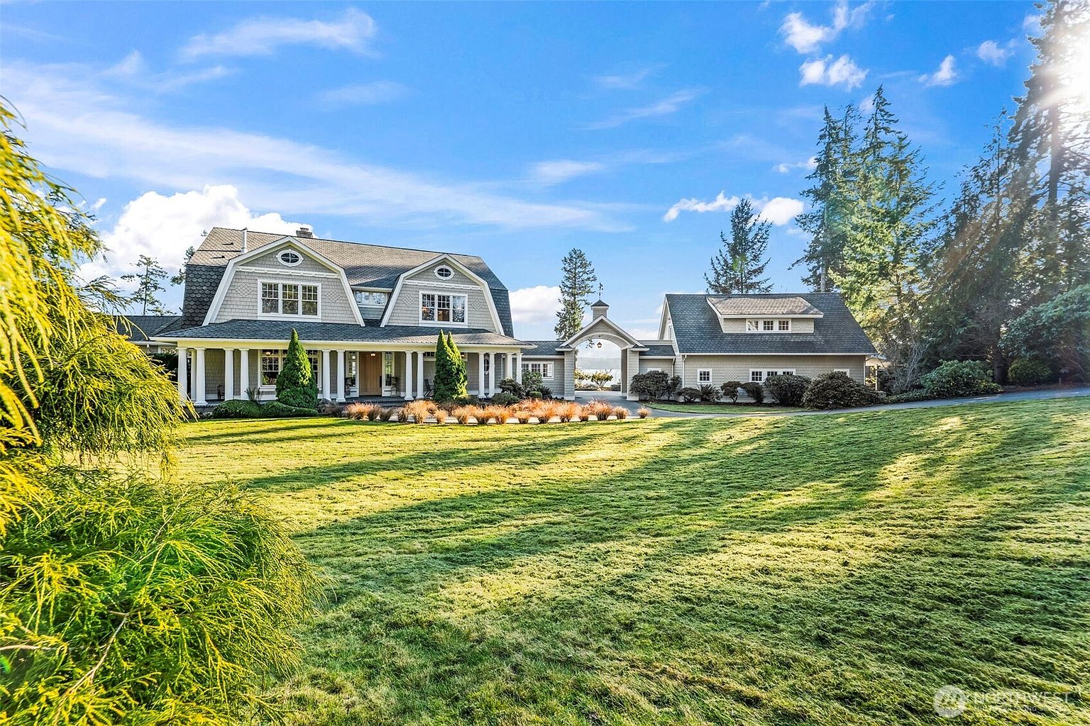 This expansive property features a traditional shingle-style home with a prominent gambrel roof and a covered front porch supported by white columns. A detached garage structure is connected to the main house via a charming arched breezeway, all set against a backdrop of mature trees and a vast, manicured lawn. The scene is captured from a low-angle perspective on a sunny day, emphasizing the grand scale and serene, park-like setting of the estate.