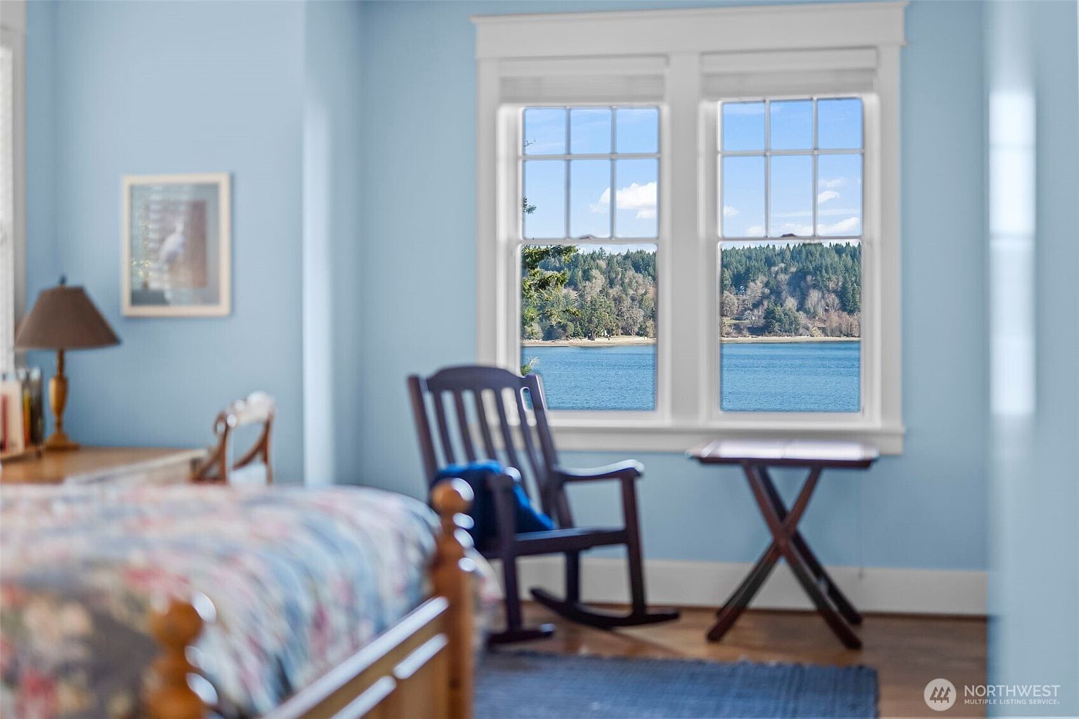 This serene bedroom features light blue walls and a view of a tranquil lake through a pair of large, white-framed windows. A wooden rocking chair sits near a small side table, creating a peaceful reading nook, while a portion of a bed with a patterned quilt is visible in the foreground. The room is bathed in natural light, emphasizing a calm and inviting atmosphere.