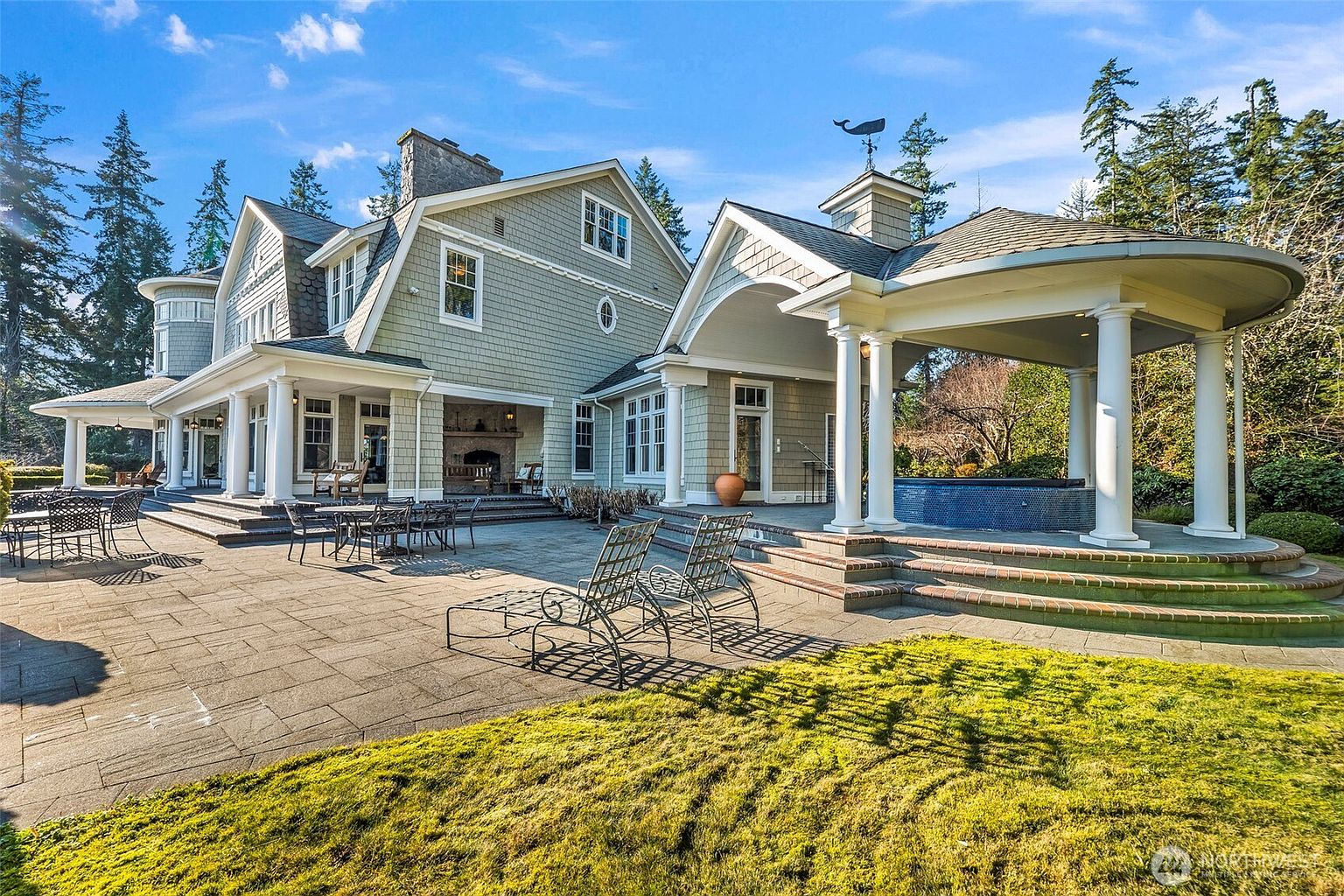 This expansive stone patio features a covered outdoor living area with a stone-clad chimney breast and a separate gazebo structure housing a circular spa. The home showcases classic shingle-style architecture with multiple white columns, large windows, and a well-manicured lawn in the foreground. The scene is captured from a low-angle perspective, emphasizing the transition between the structured outdoor living spaces and the surrounding wooded landscape.