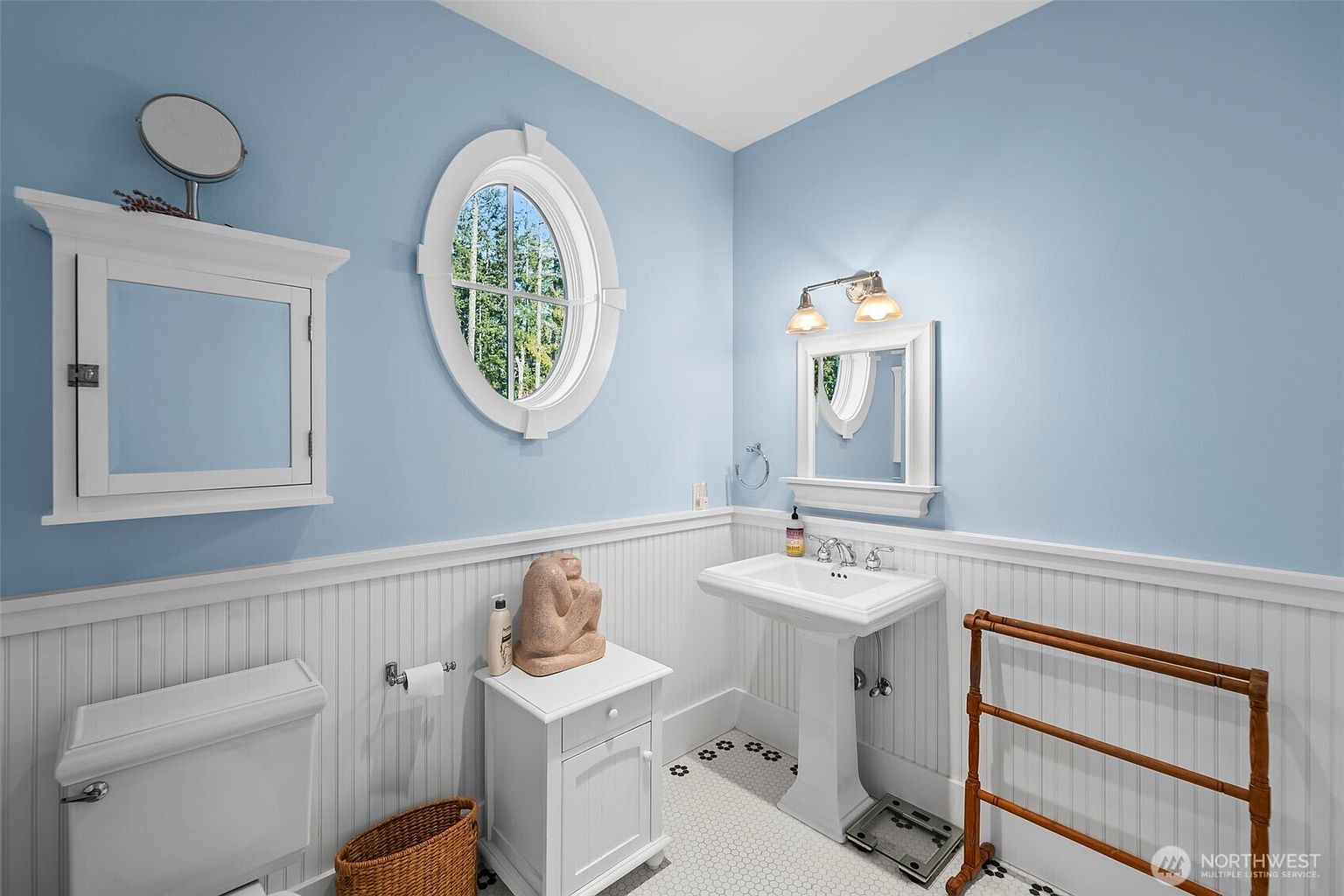 This bright, light-blue bathroom features a classic pedestal sink, a white medicine cabinet, and a matching storage cabinet topped with a decorative sculpture. The room is accented with white beadboard wainscoting, a unique oval window, and a patterned tile floor. A wooden towel rack stands to the right, completing the charming, traditional aesthetic of the space.