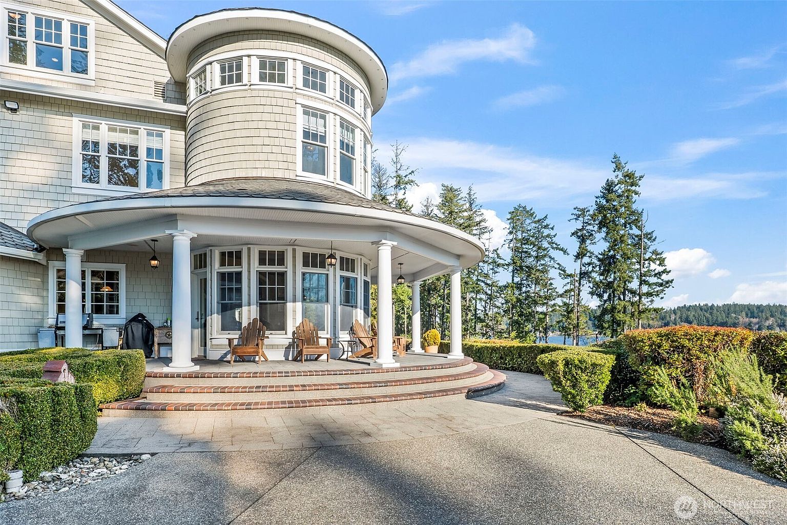 This image captures a spacious, curved covered patio attached to a shingled home with a distinctive rounded architectural tower. The patio features a paved stone surface, white columns, and a set of wooden Adirondack chairs, creating a relaxing outdoor living space. The scene is framed by manicured hedges and mature trees, with a glimpse of a body of water in the distance under a clear blue sky.