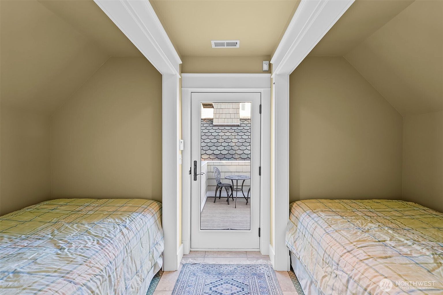 This cozy bedroom features a symmetrical layout with two twin beds positioned on either side of a central glass-paned door that leads to an outdoor balcony. The room is finished in a neutral tan paint with sloped ceilings that follow the roofline, creating an intimate, tucked-away atmosphere. A patterned area rug centers the space, while the balcony beyond provides a view of shingled exterior siding and a small bistro set.