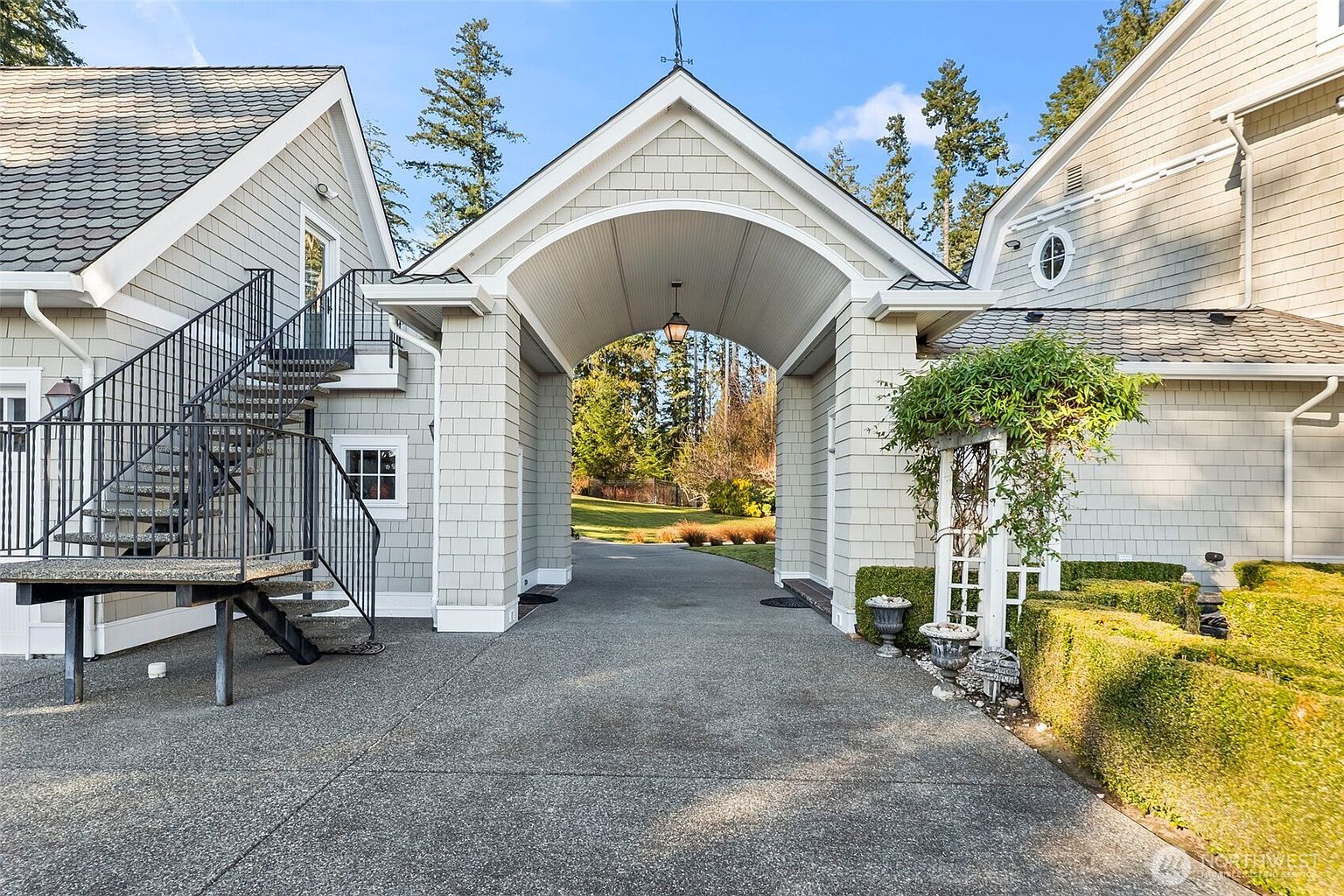 This image captures a charming porte-cochère connecting two sections of a shingled home, creating a covered driveway passage. To the left, a black metal spiral staircase leads to an upper level, while to the right, a white wooden trellis adorned with climbing greenery sits beside a manicured hedge. The perspective is centered through the archway, highlighting the architectural symmetry and the transition from the paved driveway to the lush green landscape beyond.