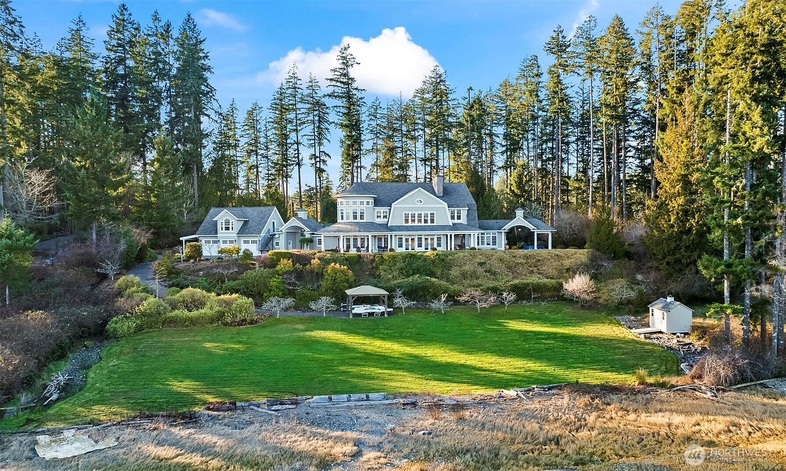 This expansive rear view captures a large, multi-level traditional home nestled against a dense backdrop of tall evergreen trees. The property features a sprawling green lawn, a small gazebo, and a detached shed, all leading down to a natural shoreline. The house itself showcases a classic architectural style with a covered porch, multiple gables, and a detached garage wing, creating a serene and private coastal estate atmosphere.