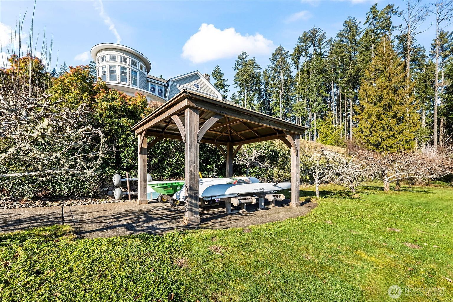 This outdoor space features a sturdy wooden gazebo structure housing a small boat on a trailer, set against a backdrop of lush green lawn and a dense forest. In the background, a multi-story home with a distinctive circular turret design rises above a landscaped slope. The scene captures a serene, private setting with a mix of manicured grass and natural woodland elements.