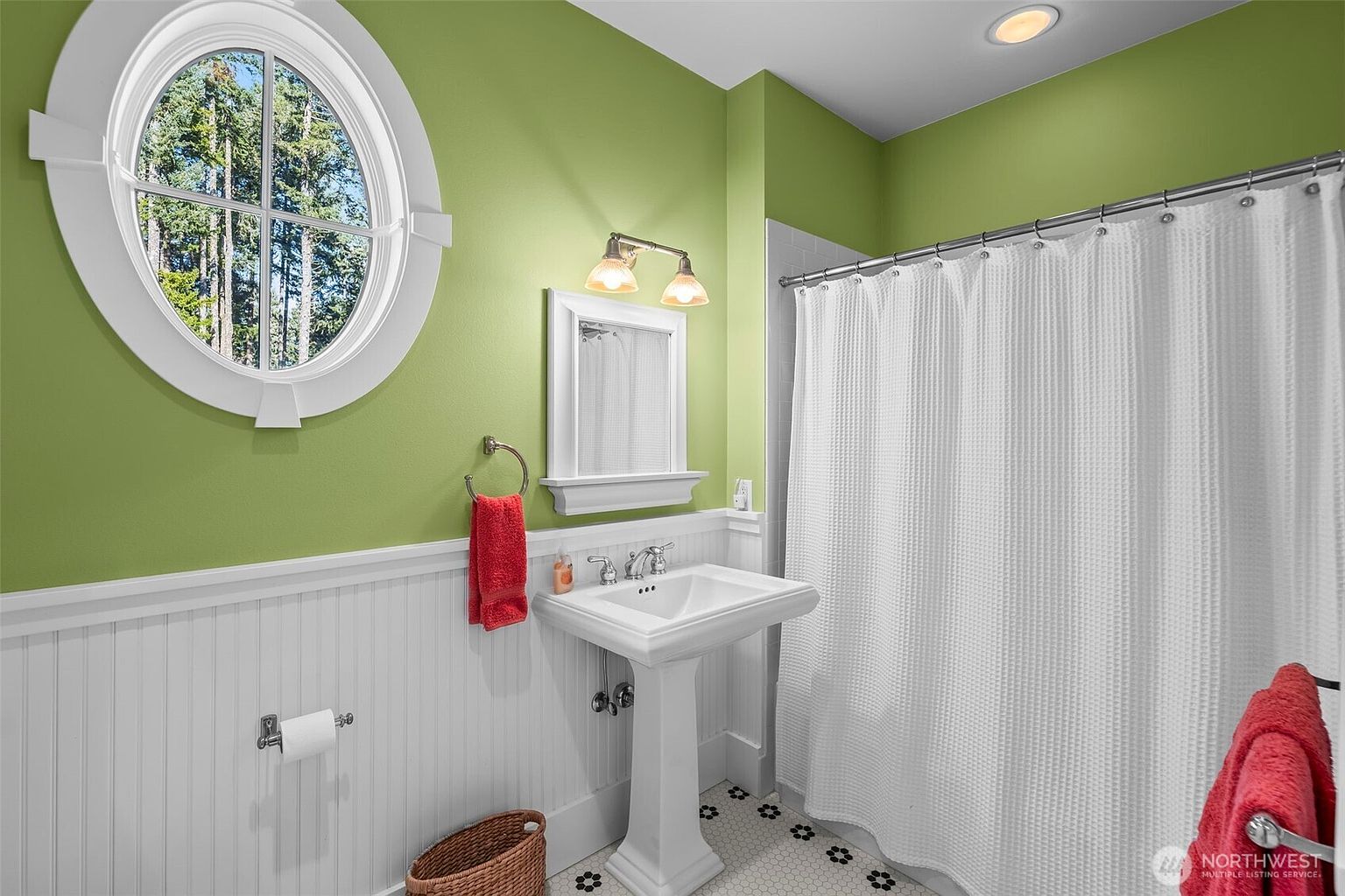 This bright bathroom features a pedestal sink, a white shower curtain, and a distinctive oval window that allows natural light to filter in. The walls are painted a vibrant green above white wainscoting, complemented by a patterned tile floor. A red towel adds a pop of color to the space, which is illuminated by a dual-bulb vanity light fixture.