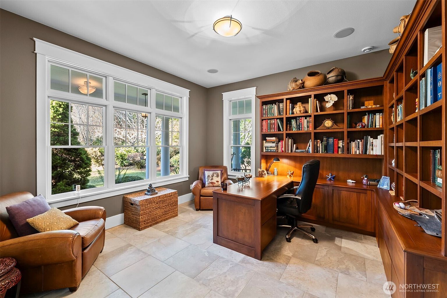This sophisticated home office features rich wood built-in shelving and a matching desk, creating a warm and professional atmosphere. Large windows with white trim allow ample natural light to fill the room, highlighting the neutral tile flooring and comfortable leather seating. The space is well-organized with a dedicated workspace and additional lounge area, making it an ideal environment for productivity.
