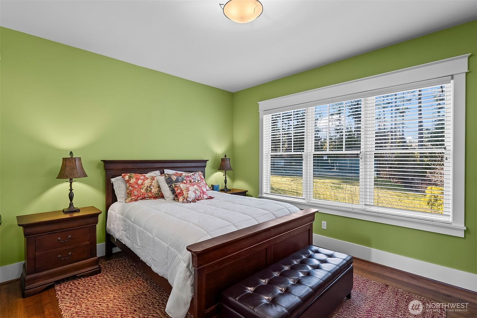 This bedroom features vibrant green walls and a dark wood bed frame dressed in white bedding with floral accent pillows. A matching dark wood nightstand with a lamp sits to the left, while a tufted leather bench is positioned at the foot of the bed. Large windows with white blinds allow natural light to fill the room, which is anchored by a textured area rug over hardwood floors.