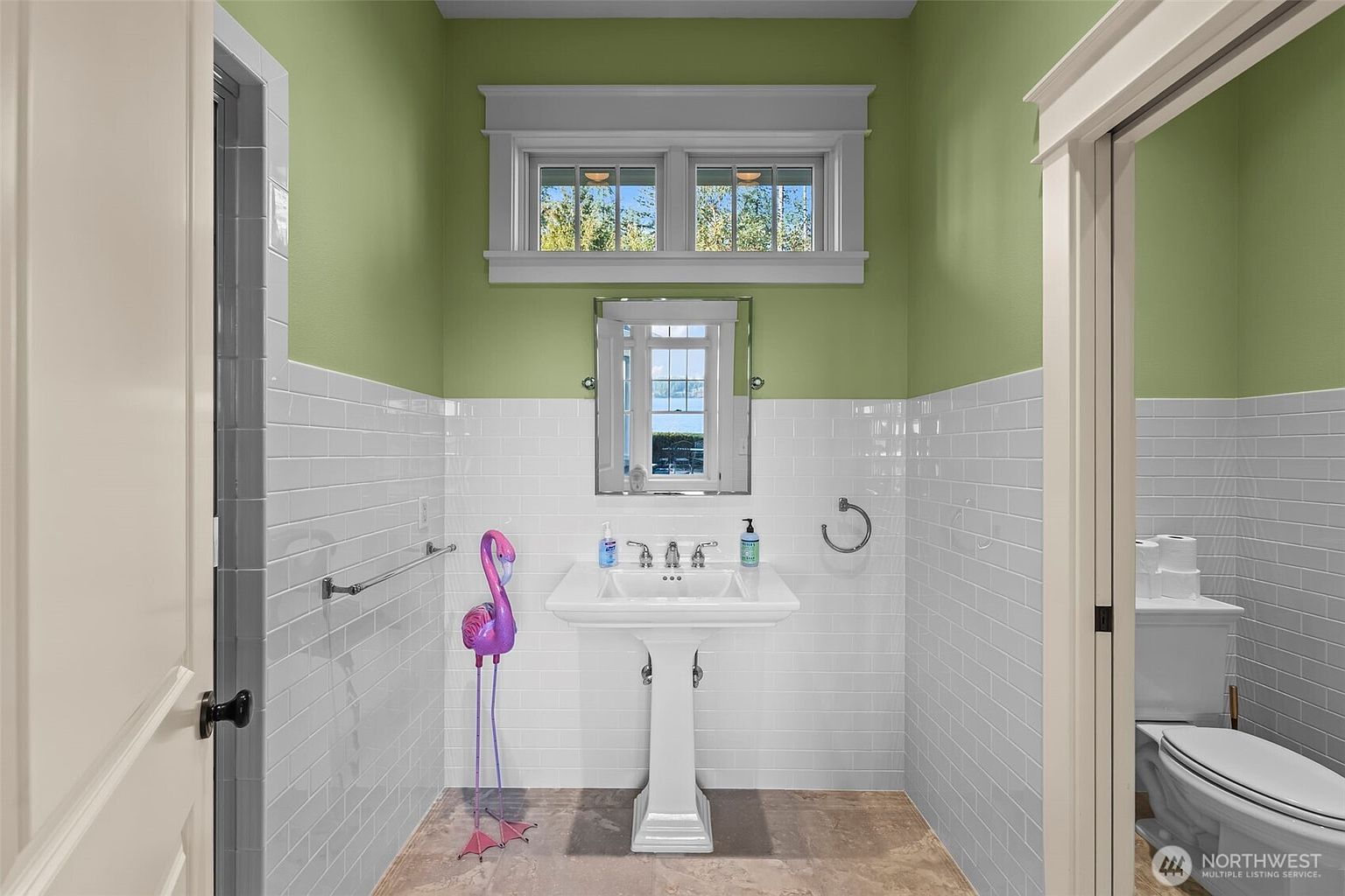 This bright powder room features a pedestal sink centered against a wall clad in white subway tile, topped with a vibrant green painted upper wall. A rectangular mirror hangs above the sink, reflecting a window that brings in natural light, while a whimsical flamingo statue adds a playful touch to the corner. The space is framed by a doorway leading to a toilet area, creating a clean and compact layout.