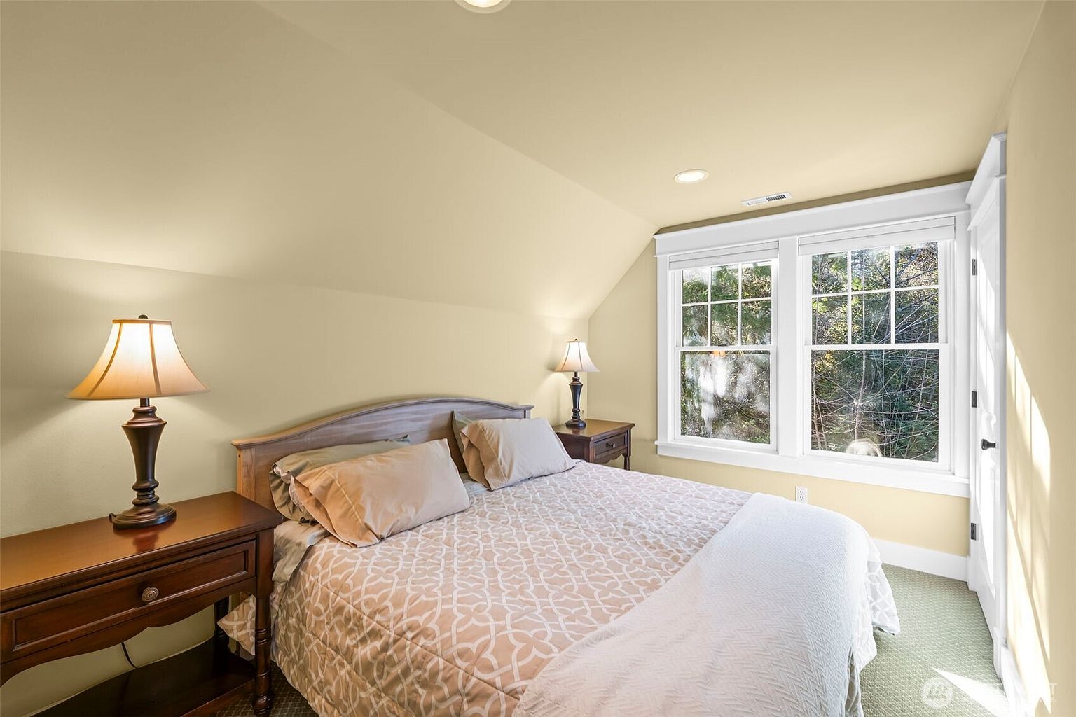 This cozy bedroom features a sloped ceiling, creating an intimate and comfortable atmosphere. The room is furnished with a bed centered between two wooden nightstands, each topped with a matching lamp, and large windows that allow natural light to brighten the space. The neutral wall color and simple decor provide a clean, inviting aesthetic suitable for a guest room.