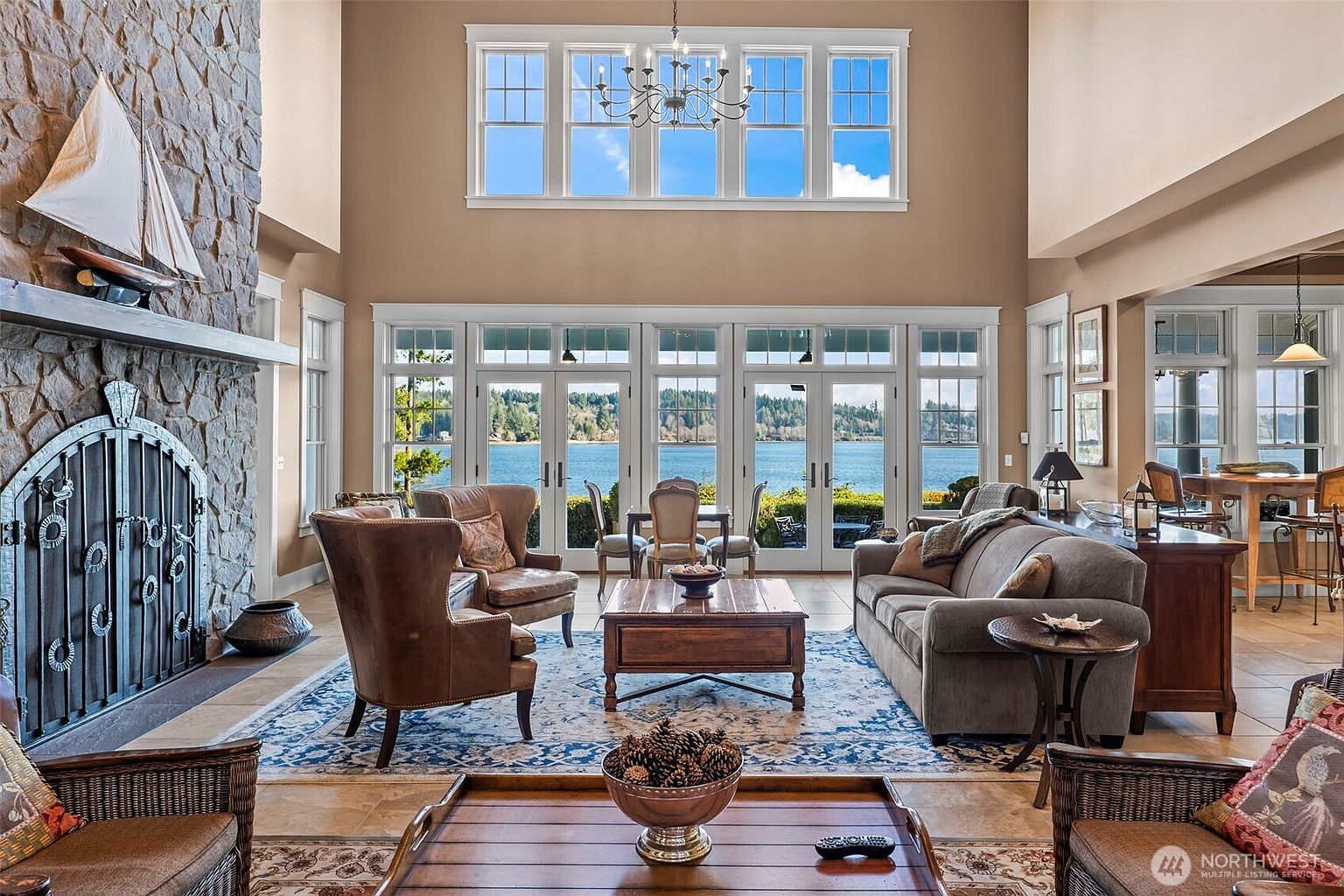 This expansive living room features a dramatic stone-clad chimney breast and a large, multi-paned window wall that frames a serene waterfront view. The space is furnished with comfortable leather armchairs and a plush sofa arranged around a central wooden coffee table, all set upon a large patterned area rug. The high ceilings and abundant natural light create an airy, sophisticated atmosphere perfect for relaxing while overlooking the water.