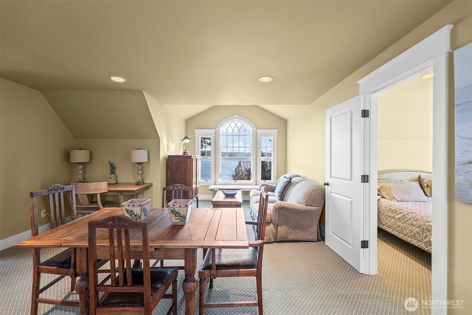 This multi-functional room features a dining area in the foreground with a wooden table and chairs, transitioning into a cozy seating area with a sofa. A large, arched window serves as a focal point, offering a scenic view of the water and surrounding trees. The space is finished with neutral-toned walls and carpet, creating a warm and inviting atmosphere that flows into an adjacent bedroom.