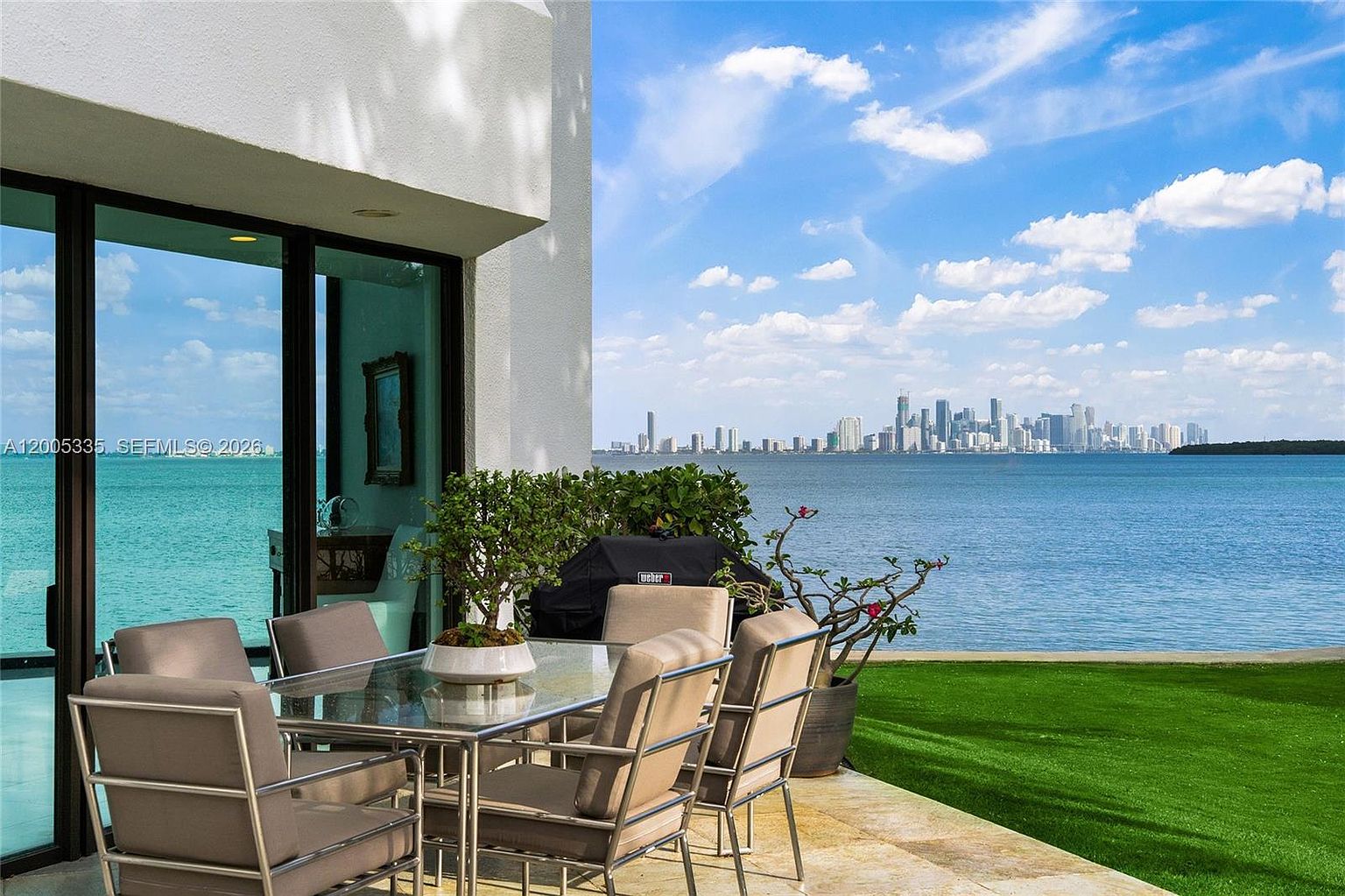 This outdoor patio space features a glass-topped dining set and a grill, positioned directly against a wall with large sliding glass doors. The area opens onto a lush green lawn that leads to a waterfront view of a distant city skyline under a bright, cloudy sky. The perspective is from a ground-level vantage point, emphasizing the seamless transition between the private patio and the expansive coastal scenery.
