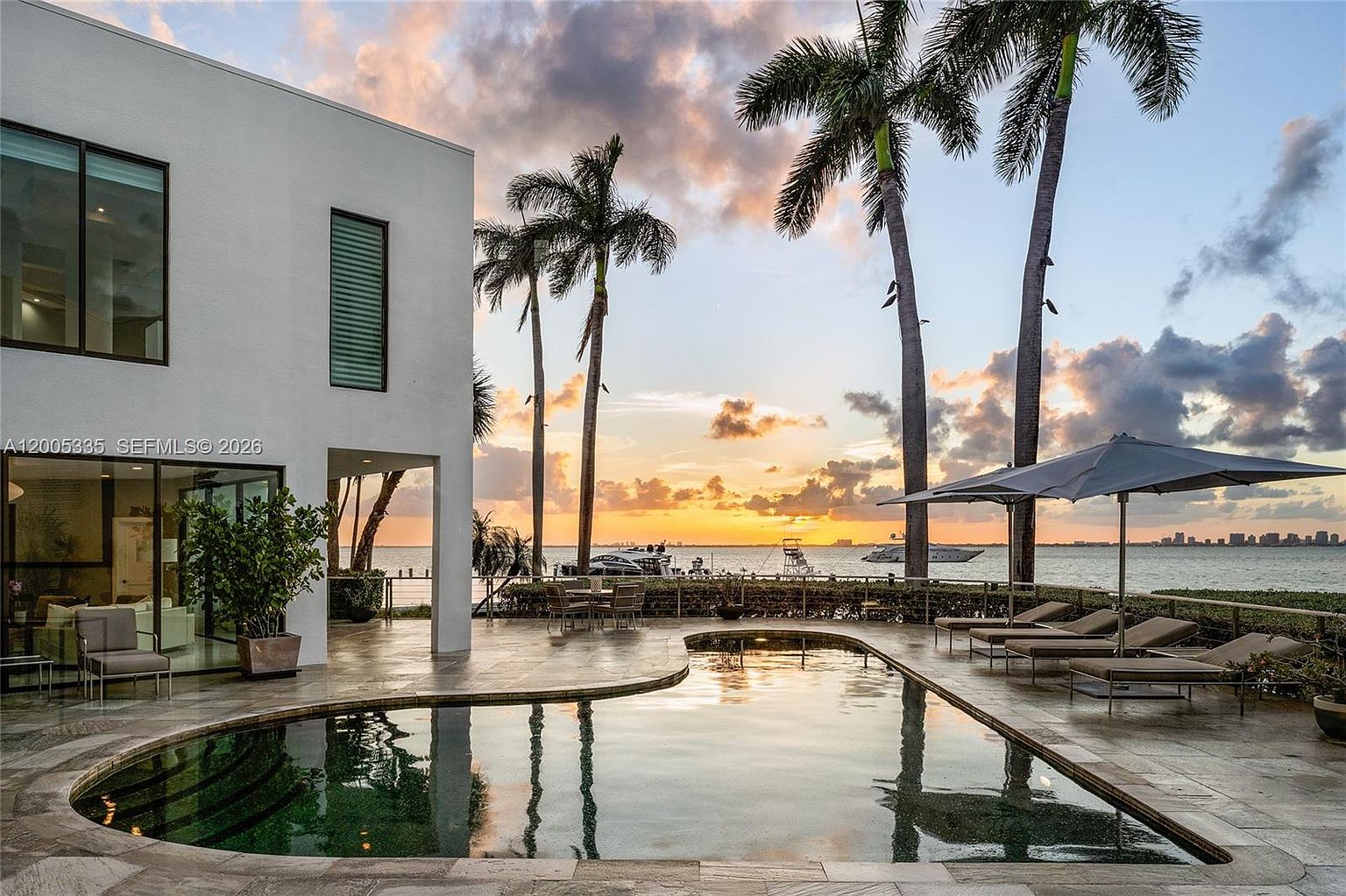 This stunning waterfront property features a curved swimming pool set against a backdrop of a vibrant sunset over the bay. The modern white exterior of the home includes large glass panels that offer a seamless transition to the outdoor patio, which is furnished with lounge chairs and an umbrella. Tall palm trees frame the horizon, creating a serene and luxurious atmosphere perfect for outdoor entertaining.