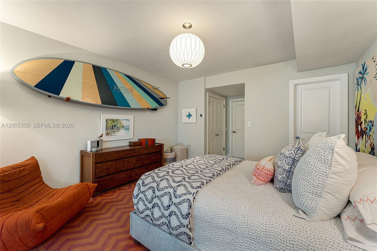 This bright and coastal-themed bedroom features a bed with patterned bedding, a vibrant orange lounge chair, and a surfboard mounted on the wall as a decorative focal point. A wooden dresser sits below a framed beach print, while a spherical pendant light hangs from the ceiling to provide soft illumination. The room is finished with light-colored walls and a textured, chevron-patterned rug, creating a relaxed and inviting atmosphere.