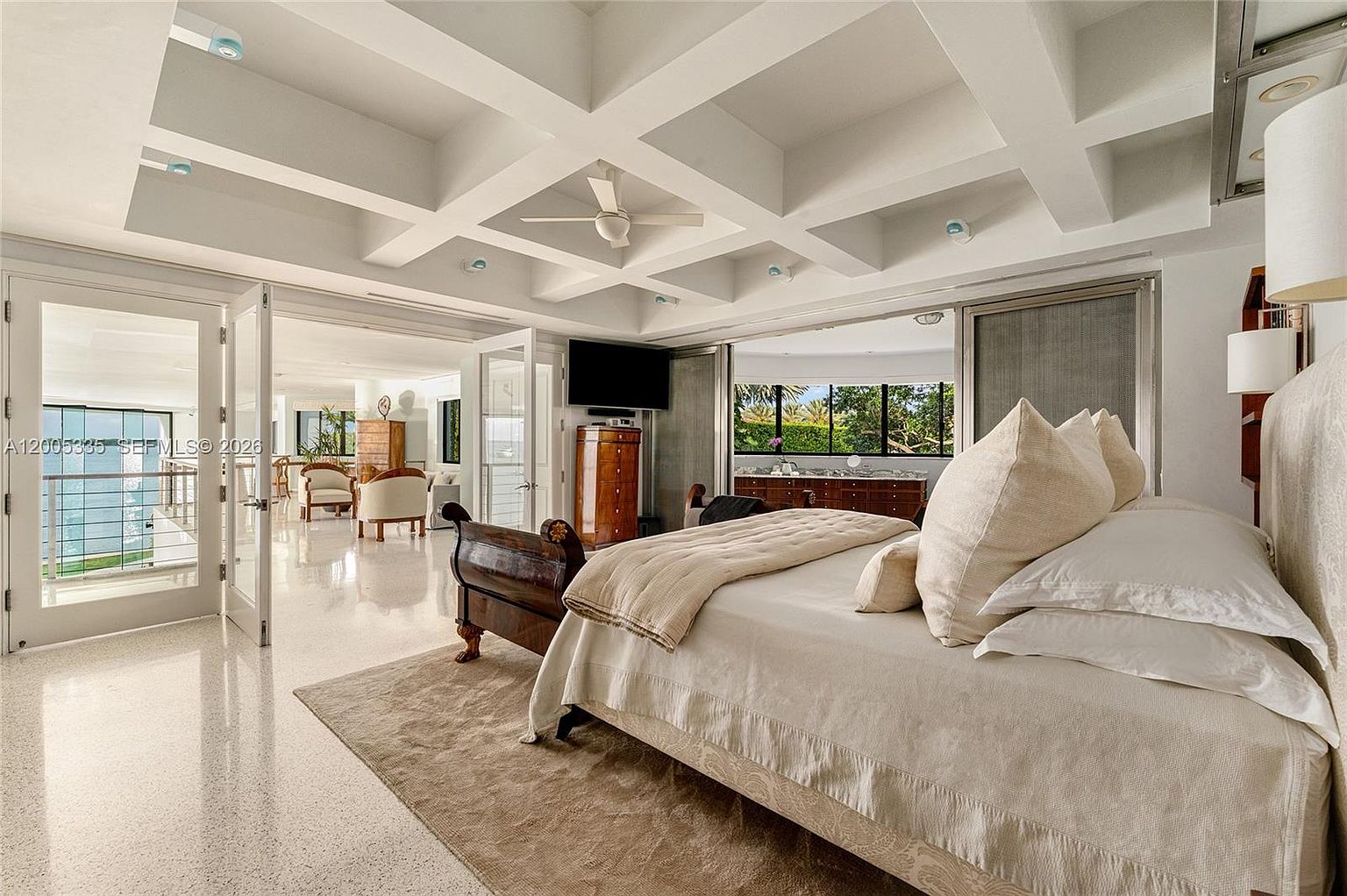 This spacious primary bedroom features a coffered ceiling with a central fan and light-colored terrazzo flooring. A large bed with neutral bedding sits on a textured area rug, while glass doors open to a balcony, providing a seamless transition to the outdoors. The room is bright and airy, with a view into an adjacent living area and large windows that allow for ample natural light.