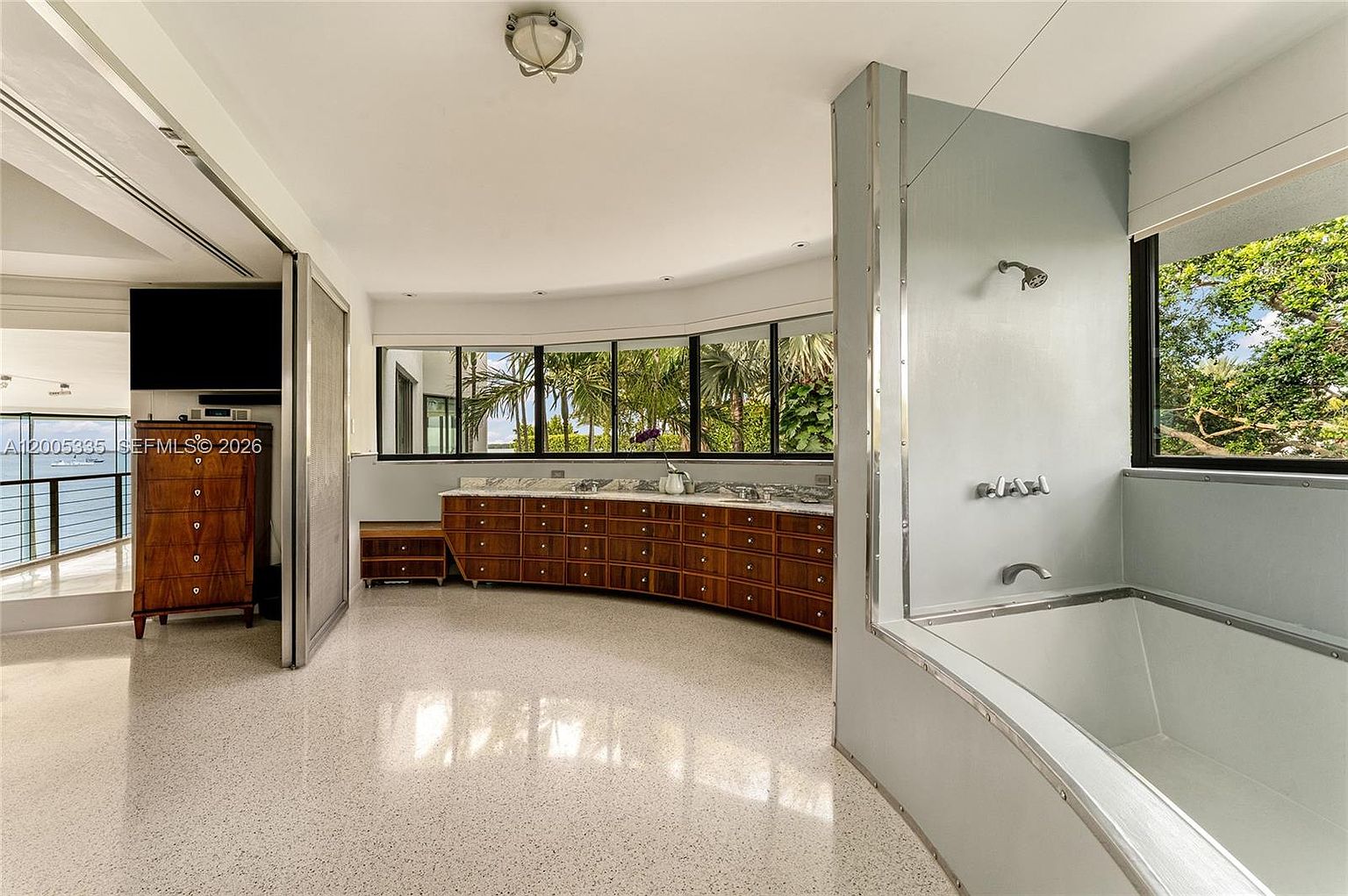This spacious primary bathroom features a large, curved vanity with extensive wood cabinetry and a stone countertop, positioned beneath a series of windows that offer a view of the surrounding greenery. To the right, a built-in bathtub is set against a light-colored wall, creating a clean and minimalist aesthetic. The room is finished with light-colored speckled flooring, providing a bright and airy atmosphere with a unique architectural layout.