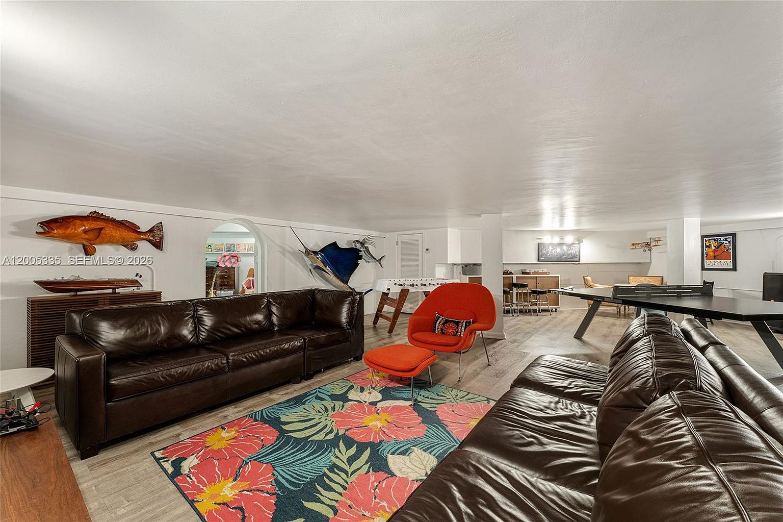 This spacious, open-concept basement features a large seating area with two dark leather sofas and a vibrant floral area rug. The room is decorated with nautical-themed wall art, including a large fish mount and a swordfish, and includes a game area with a ping pong table and a small bar setup in the background. The space has light-toned flooring and a neutral color palette, creating a versatile recreational area.