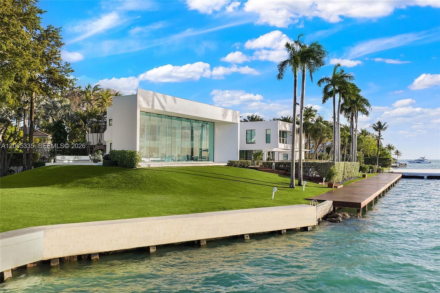 This modern waterfront estate features a striking white architectural design with expansive floor-to-ceiling glass walls that offer panoramic views of the bay. A manicured green lawn slopes gently toward a concrete seawall and a private wooden dock, creating a seamless transition between the residence and the water. Tall palm trees frame the property, enhancing the tropical, luxury aesthetic of this coastal home.