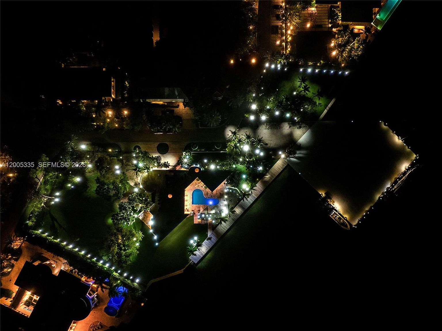 This nighttime aerial perspective captures a sprawling waterfront estate illuminated by extensive landscape lighting. The property features a central swimming pool, manicured lawns, and a long wooden dock extending into the dark water, creating a dramatic and luxurious atmosphere. The composition highlights the expansive grounds and the sophisticated outdoor lighting design, emphasizing the property's privacy and high-end appeal.