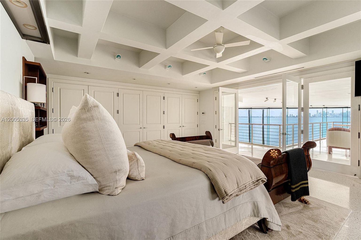 This spacious primary bedroom features a coffered ceiling with a central fan and a neutral color palette that creates a serene atmosphere. A large bed with plush bedding serves as the focal point, while a set of French doors opens onto a balcony with expansive water views. The room is finished with light-toned flooring and built-in cabinetry, offering a sophisticated and airy aesthetic.