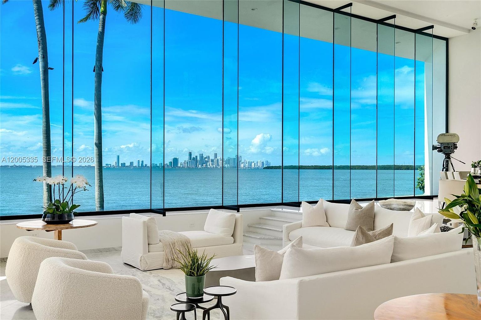 This expansive living room features a minimalist aesthetic with white plush seating arranged to face a massive wall of floor-to-ceiling glass. The glazing provides an unobstructed, panoramic view of the bay and the distant city skyline, creating a seamless connection between the interior and the water. A vintage-style film camera on a tripod stands to the right, adding a unique, artistic touch to the bright, airy space.