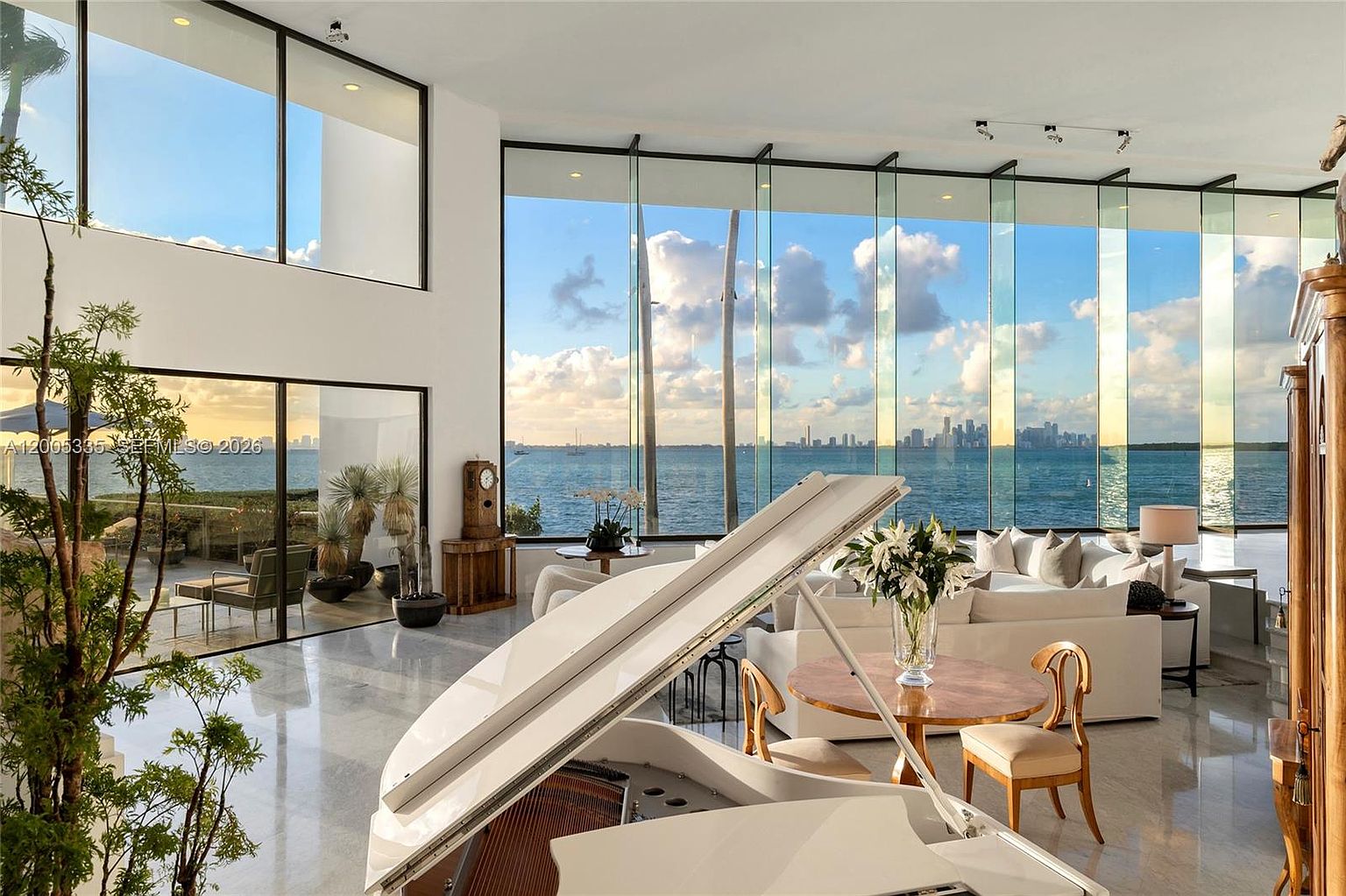 This expansive living area features a grand white piano in the foreground and a sophisticated seating arrangement centered around a circular dining table. The space is defined by massive floor-to-ceiling windows that provide an unobstructed, panoramic view of the ocean and a distant city skyline. The interior aesthetic is bright and airy, with polished light-toned flooring and a seamless transition to an outdoor terrace, creating a luxurious and serene atmosphere.