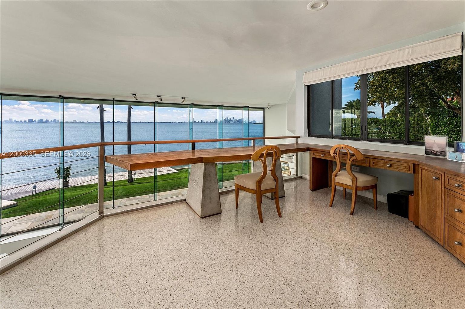 This bright home office features a long, custom-built desk with a concrete pedestal base, positioned to take full advantage of the expansive water views. The room is enclosed by floor-to-ceiling glass walls that provide a seamless connection to the outdoors, while a secondary wooden desk area with built-in cabinetry offers additional workspace. The space is finished with light-colored speckled flooring, creating a clean and professional atmosphere.