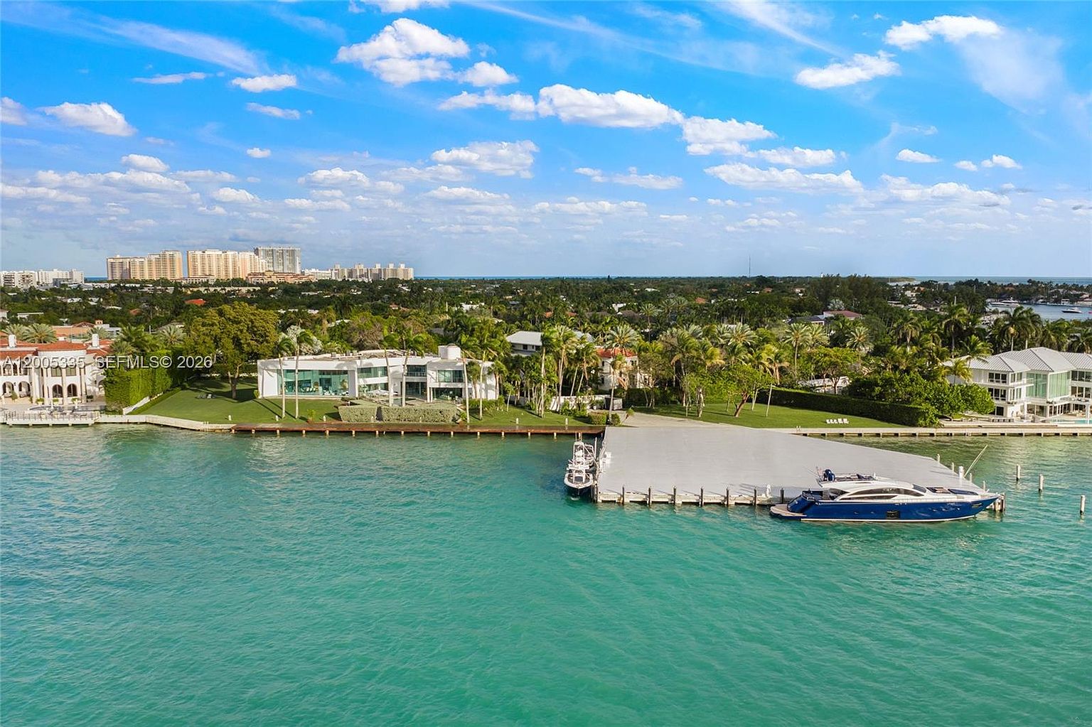 This high-angle aerial view captures a luxurious waterfront estate situated along a vibrant turquoise bay. The property features a sprawling modern residence with expansive glass walls, a manicured lawn, and a private dock accommodating a large yacht. The surrounding landscape is lush with tropical palm trees, while the distant horizon reveals a cityscape under a bright, cloud-dotted sky.
