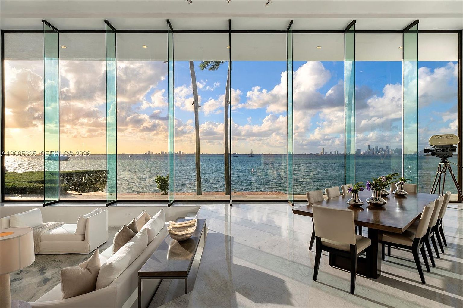 This expansive open-concept living and dining area features dramatic floor-to-ceiling glass walls that frame a panoramic view of the bay and city skyline. The space is finished with polished light-toned stone flooring and includes a contemporary dining set and a plush white sofa arrangement. A vintage-style film camera on a tripod stands in the corner, adding a unique artistic touch to the sophisticated, light-filled interior.