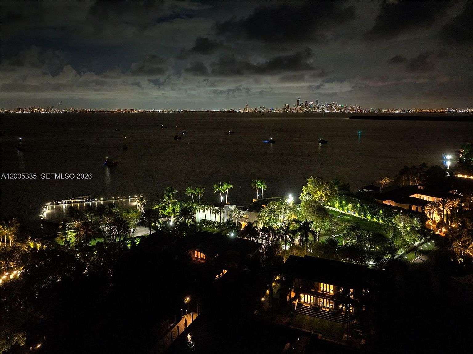 This stunning aerial night shot captures a luxurious waterfront estate overlooking a calm bay with a distant city skyline glowing on the horizon. The property features lush, illuminated landscaping, a private dock extending into the water, and multiple multi-story residences bathed in warm, inviting light. The perspective provides a cinematic overview of the expansive grounds and the serene coastal setting.