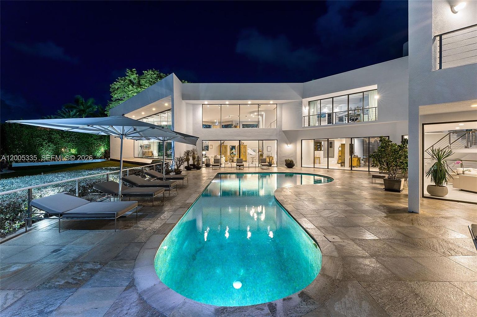 This striking modern estate features a large, curved swimming pool set against a white, multi-level architectural structure. The expansive stone-paved patio includes multiple lounge chairs under a large umbrella, creating a luxurious outdoor living space. Large glass panels and windows provide a seamless transition between the interior living areas and the illuminated pool deck at night.