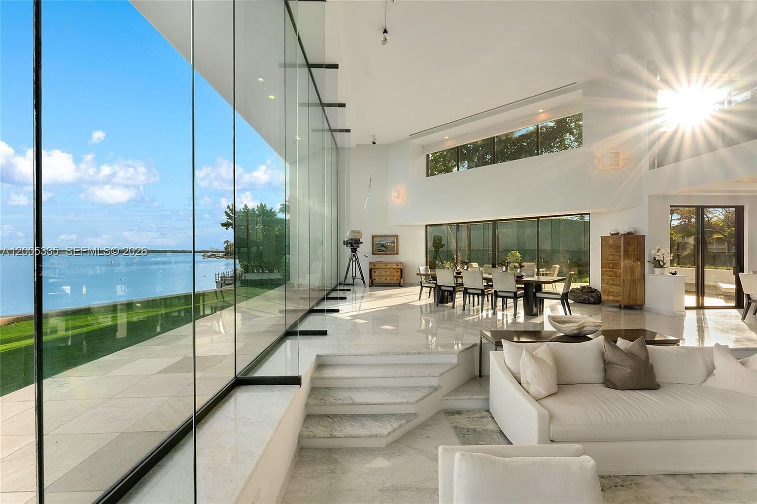 This expansive, open-concept living and dining area features dramatic floor-to-ceiling glass walls that provide an unobstructed, panoramic view of the waterfront. The space is characterized by bright, white interiors, polished stone flooring, and a sunken living room design that creates a distinct separation from the dining area. A large dining table sits centrally, while the bright, airy atmosphere is enhanced by the natural light streaming in from both the glass walls and the high-set windows.