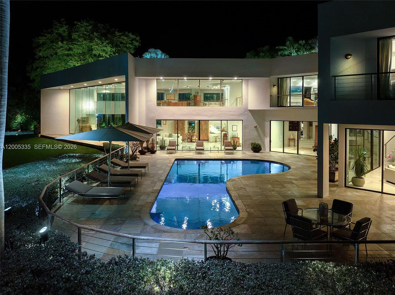 This modern, multi-level residence is captured at night, showcasing a sprawling rear patio and a custom-shaped swimming pool. The home features extensive glass walls that provide a clear view into the illuminated interior living spaces, while a tiered terrace offers ample room for outdoor lounging and dining. The scene is framed by lush landscaping and ambient exterior lighting, creating a sophisticated and inviting atmosphere for evening entertaining.