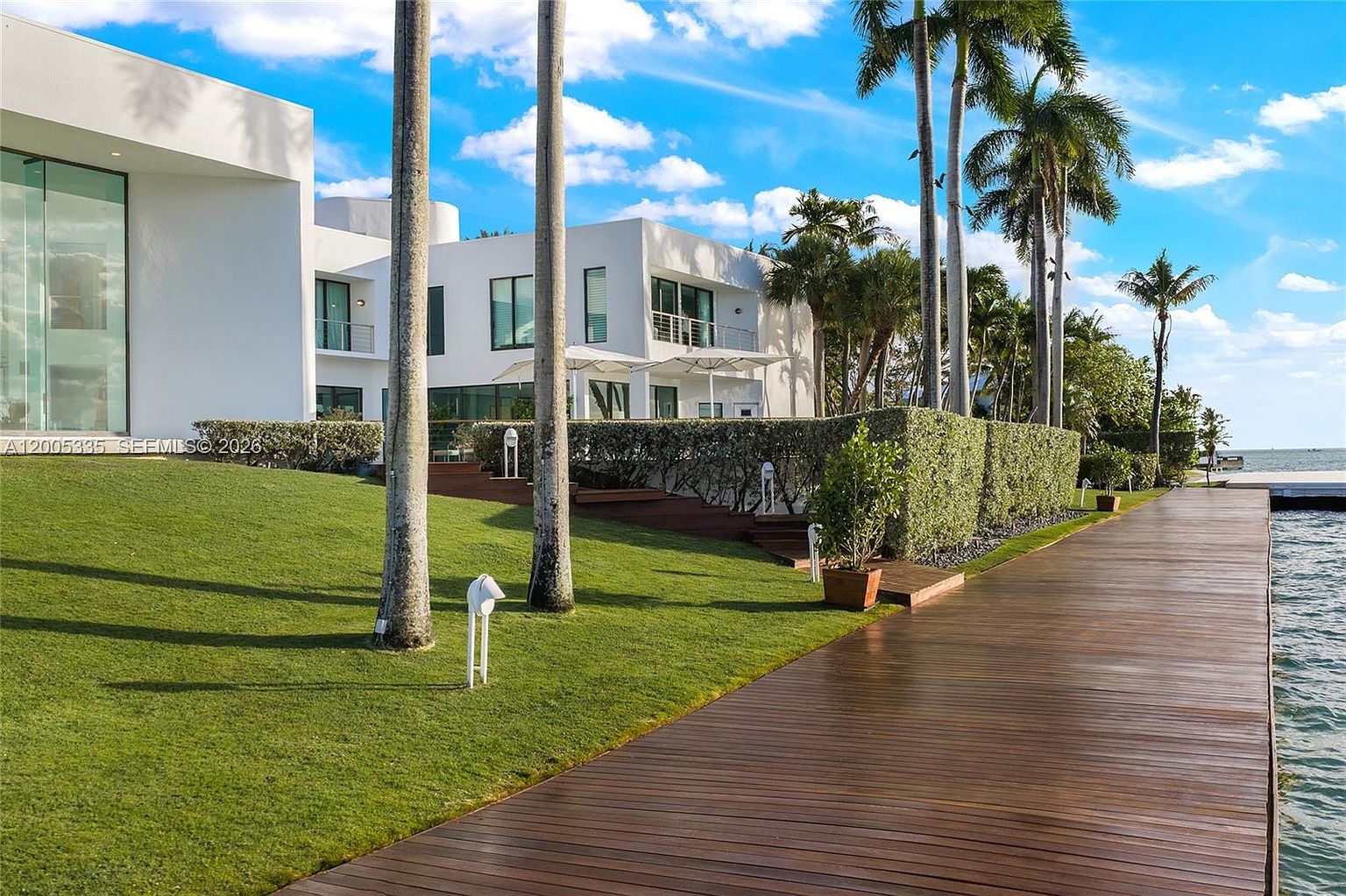 This rear view of a modern, white-stucco luxury residence showcases a sprawling lawn that transitions into a long, wooden dock extending over the water. The architecture features clean lines, large glass panels, and multiple levels, complemented by tall palm trees and manicured hedges. The perspective is captured from the dock, emphasizing the seamless connection between the home's outdoor living space and the waterfront setting.