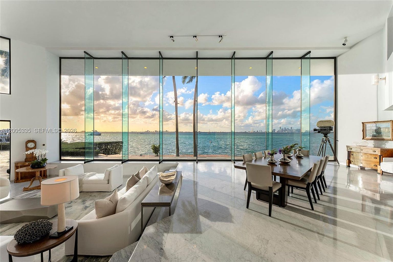 This expansive open-concept living and dining area features polished stone flooring and a dramatic wall of floor-to-ceiling glass panels that frame a panoramic waterfront view. The space is furnished with contemporary white seating and a large wooden dining table, creating a sophisticated atmosphere. A vintage-style camera on a tripod stands as a unique decorative accent near the dining area, enhancing the room's artistic and luxurious aesthetic.