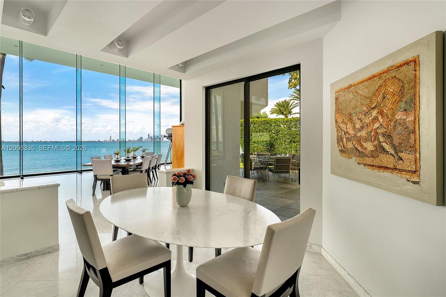 This bright dining area features a round stone-topped table surrounded by four modern, light-colored upholstered chairs. The space is defined by expansive floor-to-ceiling windows that provide a panoramic view of the ocean and city skyline, while a sliding glass door leads to an outdoor patio. A large, textured piece of art hangs on the wall, adding a sophisticated touch to the minimalist, light-filled interior.