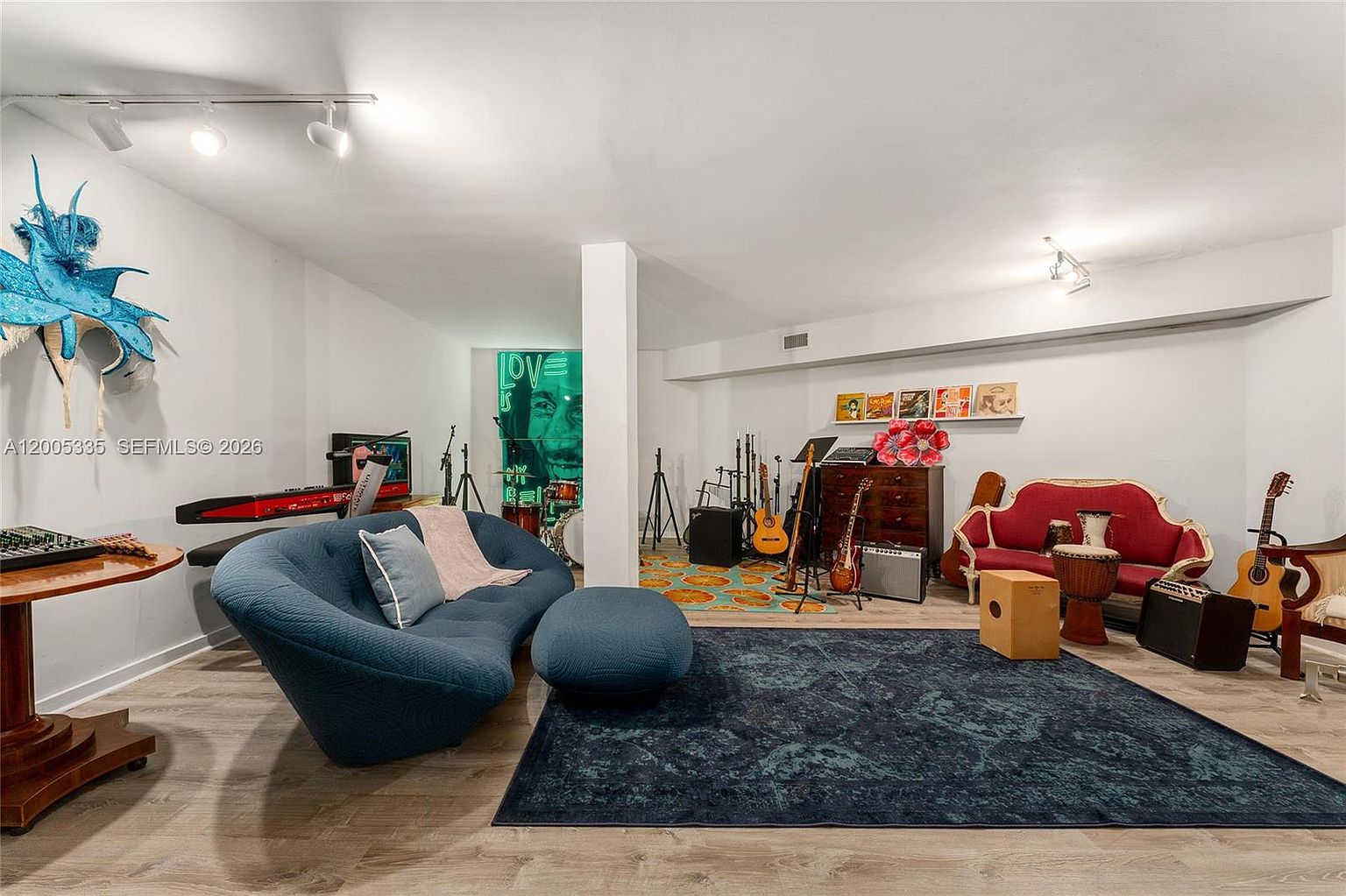 This spacious, open-concept basement serves as a creative music and lounge area, featuring light-toned wood-look flooring and a large, dark blue area rug. The room is furnished with a contemporary curved blue sofa, a matching ottoman, and a vintage-style red settee, while the background is filled with various musical instruments including guitars, a drum kit, and amplifiers. A prominent wall mural adds a vibrant artistic touch to the space, which is illuminated by track lighting and recessed ceiling lights.
