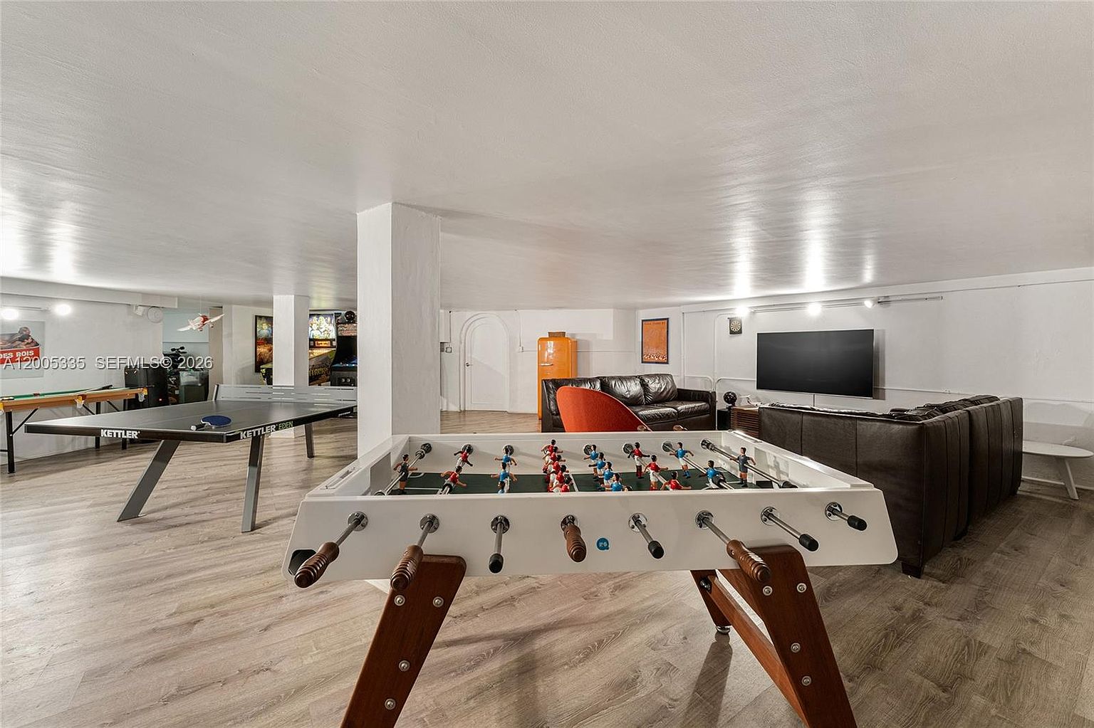 This spacious, open-concept basement serves as a recreational lounge, featuring a foosball table in the foreground and a ping-pong table to the left. The room is furnished with a large, dark leather sectional sofa and a television, creating a comfortable area for entertainment. The space is finished with light-toned wood-look flooring and white walls, providing a bright and versatile environment for leisure activities.