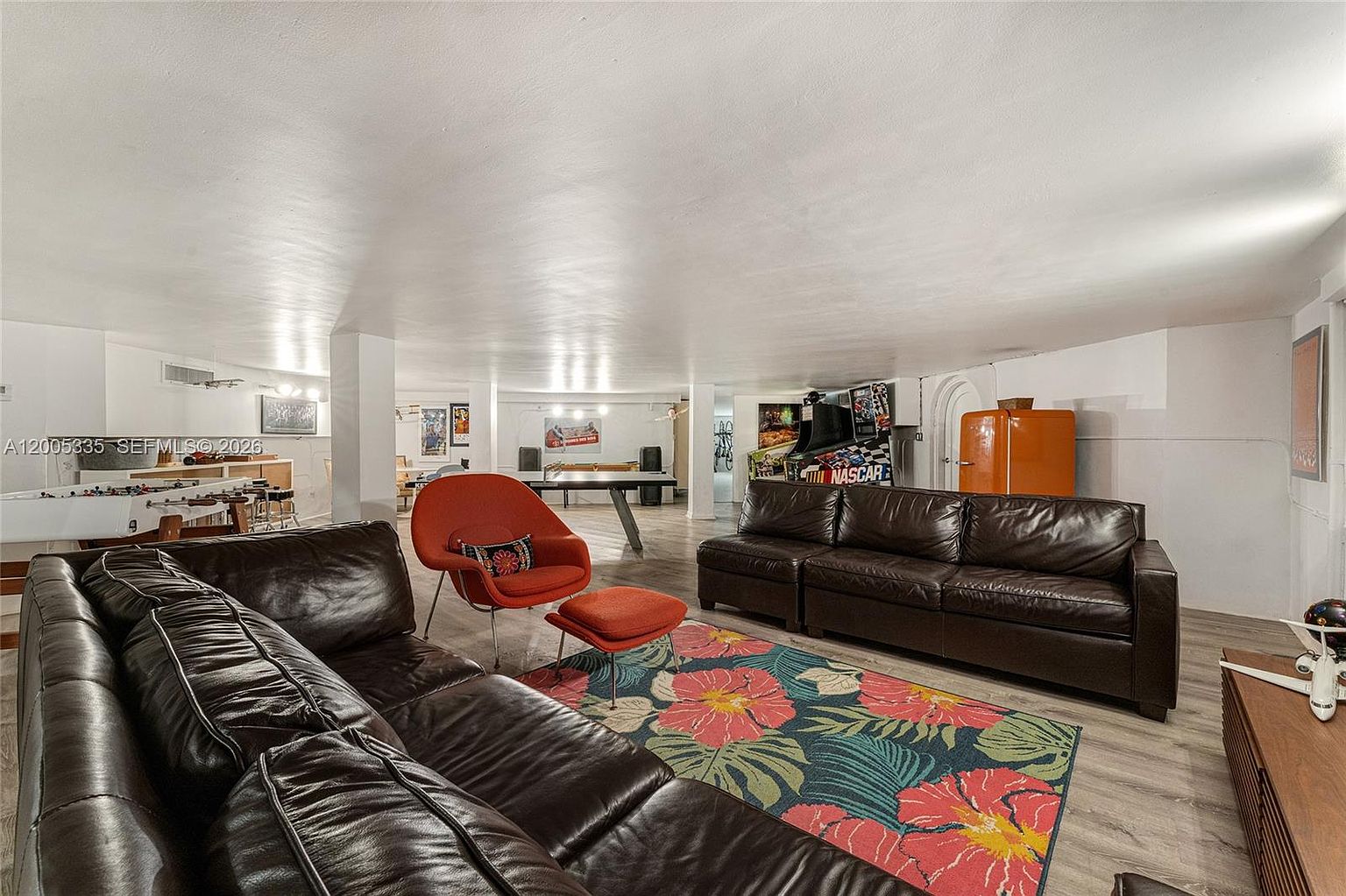 This expansive, open-concept basement serves as a multi-functional recreation area, featuring a large seating arrangement with dark leather sofas and a vibrant floral area rug. The space is equipped with various entertainment options, including a foosball table, a ping pong table, and a vintage-style arcade game. The room is illuminated by recessed lighting and maintains a casual, retro-inspired aesthetic with a bright orange refrigerator and mid-century modern accent chair.