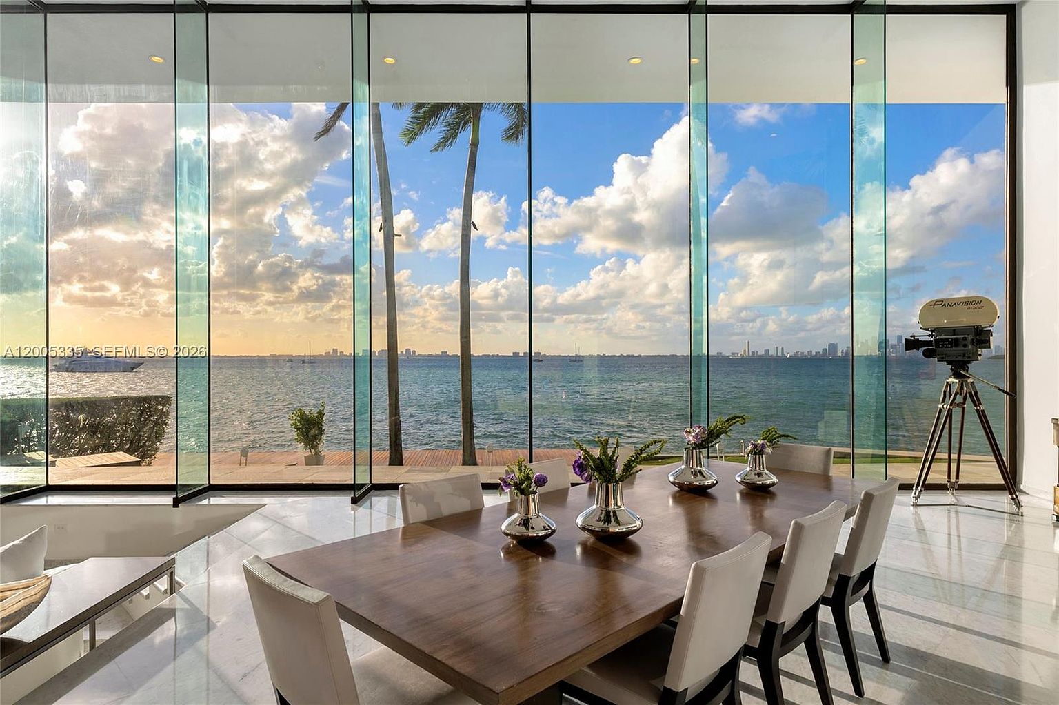 This elegant dining room features a large wooden table surrounded by light-colored upholstered chairs, set against a backdrop of expansive floor-to-ceiling glass walls. The room offers a panoramic, unobstructed view of the ocean, a distant city skyline, and palm trees, creating a seamless transition between indoor and outdoor spaces. A vintage-style film camera on a tripod stands in the corner, adding a unique, artistic touch to the sophisticated, light-filled interior.