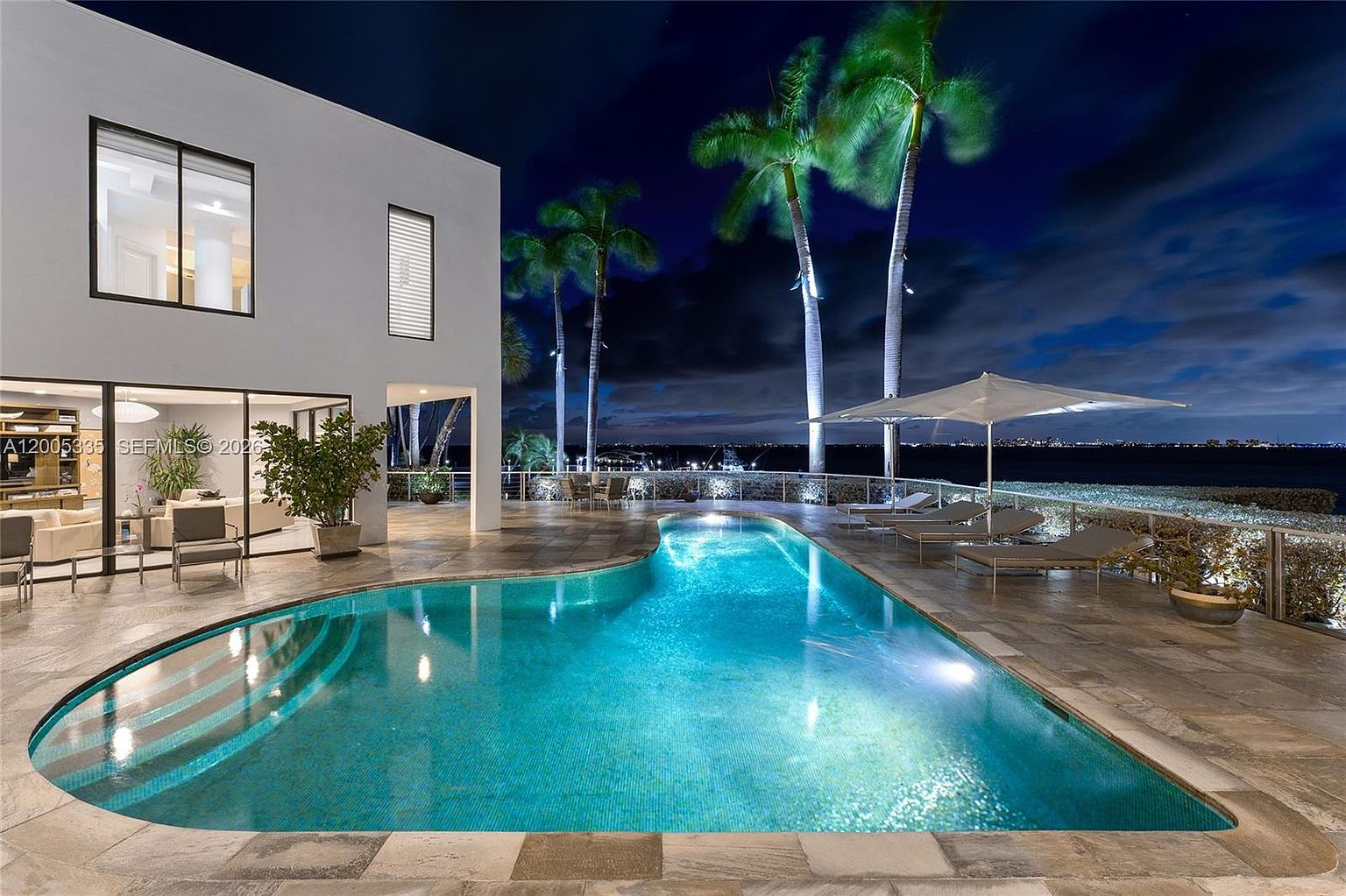 This stunning nighttime view captures a modern, curvilinear swimming pool set against a waterfront backdrop. The pool deck is paved with large stone tiles and features lounge chairs under a white umbrella, while tall palm trees frame the scene. The adjacent home showcases contemporary architecture with large glass sliding doors that open onto the patio, creating a seamless indoor-outdoor living experience.