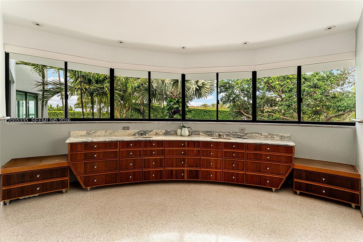 This spacious bathroom features a long, curved vanity with extensive wood cabinetry and a light-colored stone countertop with dual sinks. A series of large windows follows the curve of the wall, providing an expansive view of the lush greenery outside and filling the room with natural light. The space is finished with a neutral, speckled floor, creating a bright and airy atmosphere.