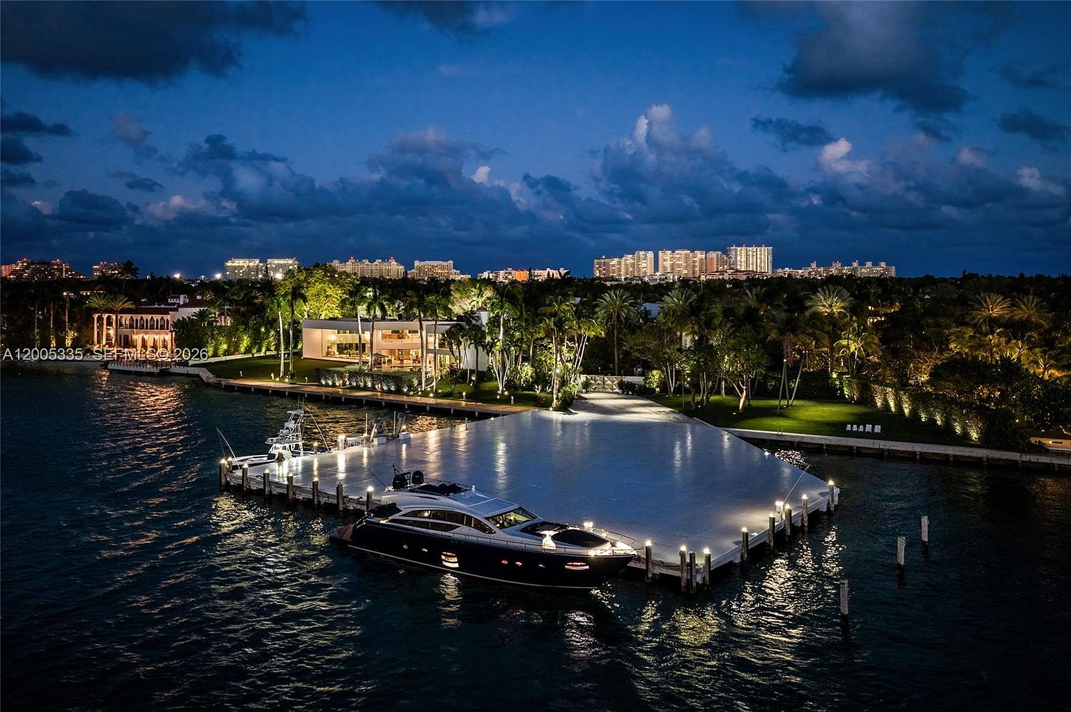 This stunning aerial night shot captures a luxurious waterfront estate featuring a large, illuminated private dock with a sleek yacht moored alongside. The property is surrounded by lush, manicured landscaping and palm trees, with a modern residence glowing warmly in the background. The scene is set against a dramatic twilight sky, with the distant city skyline providing a sophisticated urban backdrop.