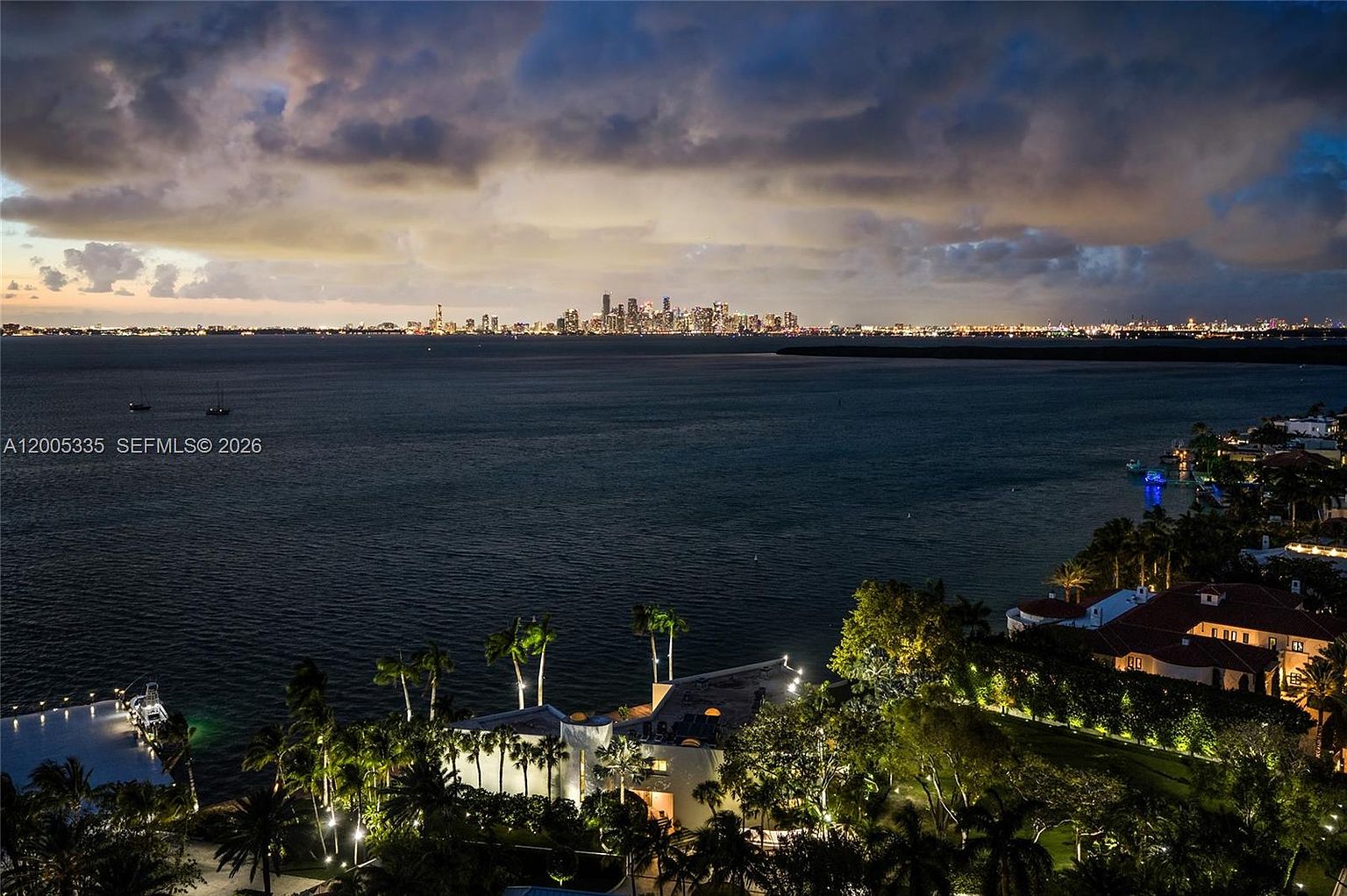 This high-angle aerial view captures a luxurious waterfront estate at dusk, set against the backdrop of a distant, illuminated city skyline across the bay. The property features a sprawling residence with multiple lit structures, lush tropical landscaping, and a private dock extending into the calm water. The dramatic, cloud-filled sky and the contrast between the warm residential lighting and the cool blue tones of the evening create a sophisticated and serene atmosphere.