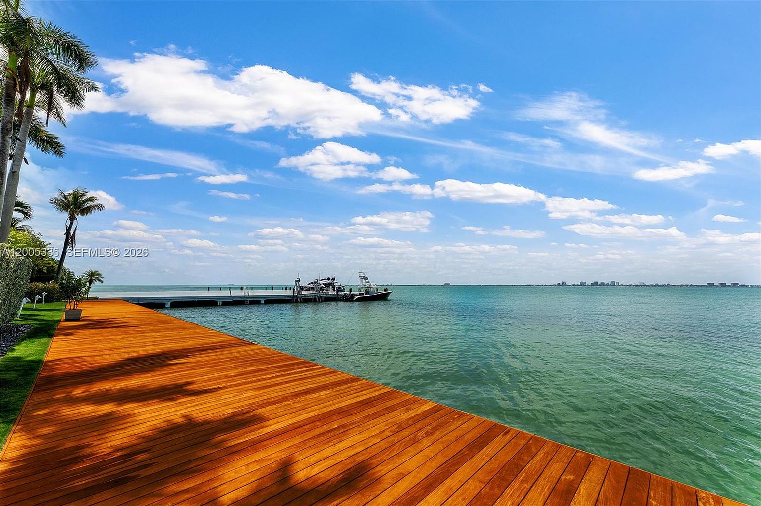 This image captures a wide, polished wooden deck extending toward a serene, expansive bay. A long private dock stretches out into the calm turquoise water, where a boat is moored under a bright, partly cloudy sky. The perspective is low and wide, emphasizing the seamless transition from the luxury outdoor living space to the open waterfront.